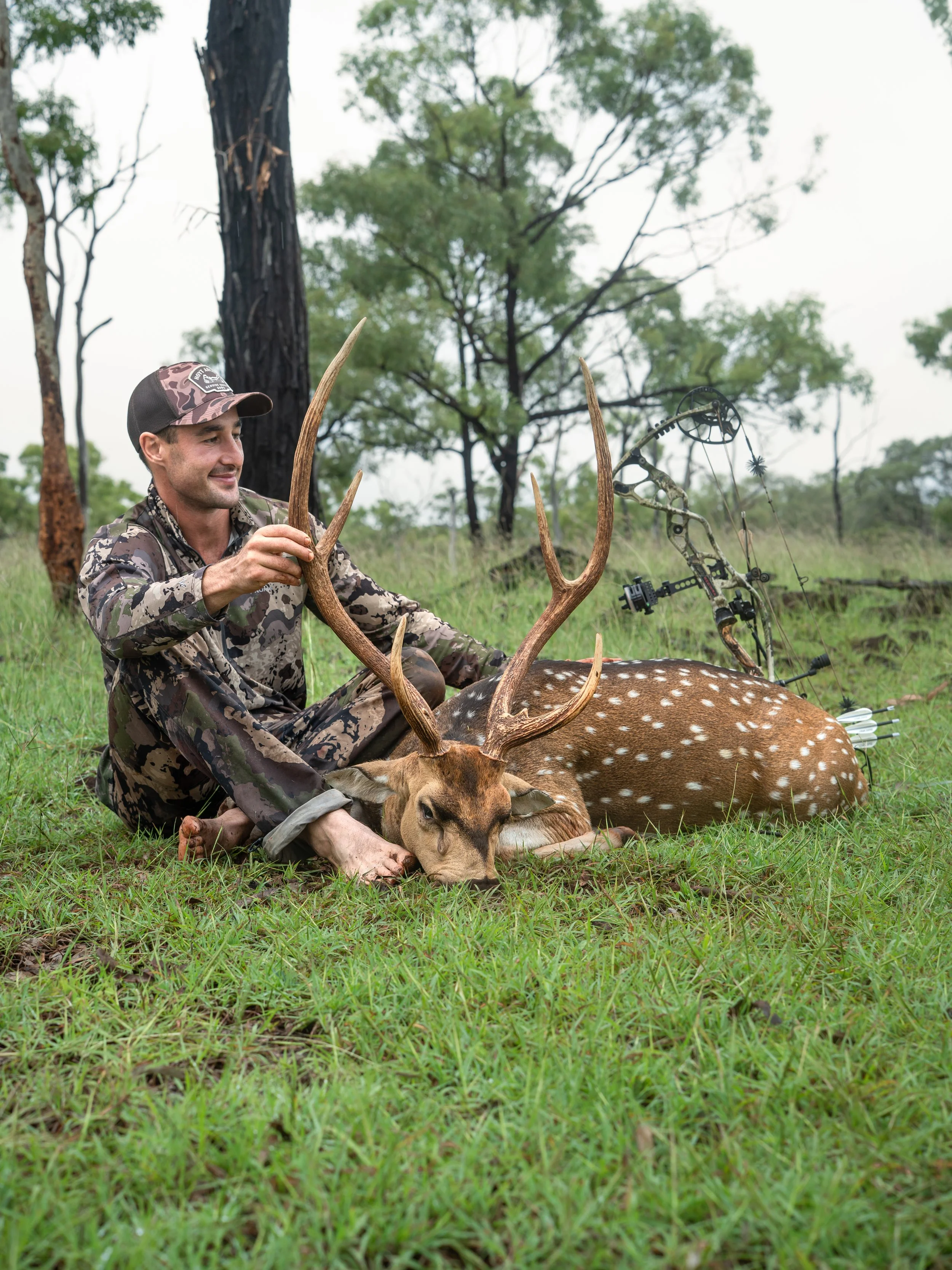 A man in camouflage clothing and a cap is sitting on the grass, smiling, next to a large deer with antlers, which is lying down. There is a compound bow with arrows set nearby, and the background features trees and a cloudy sky.