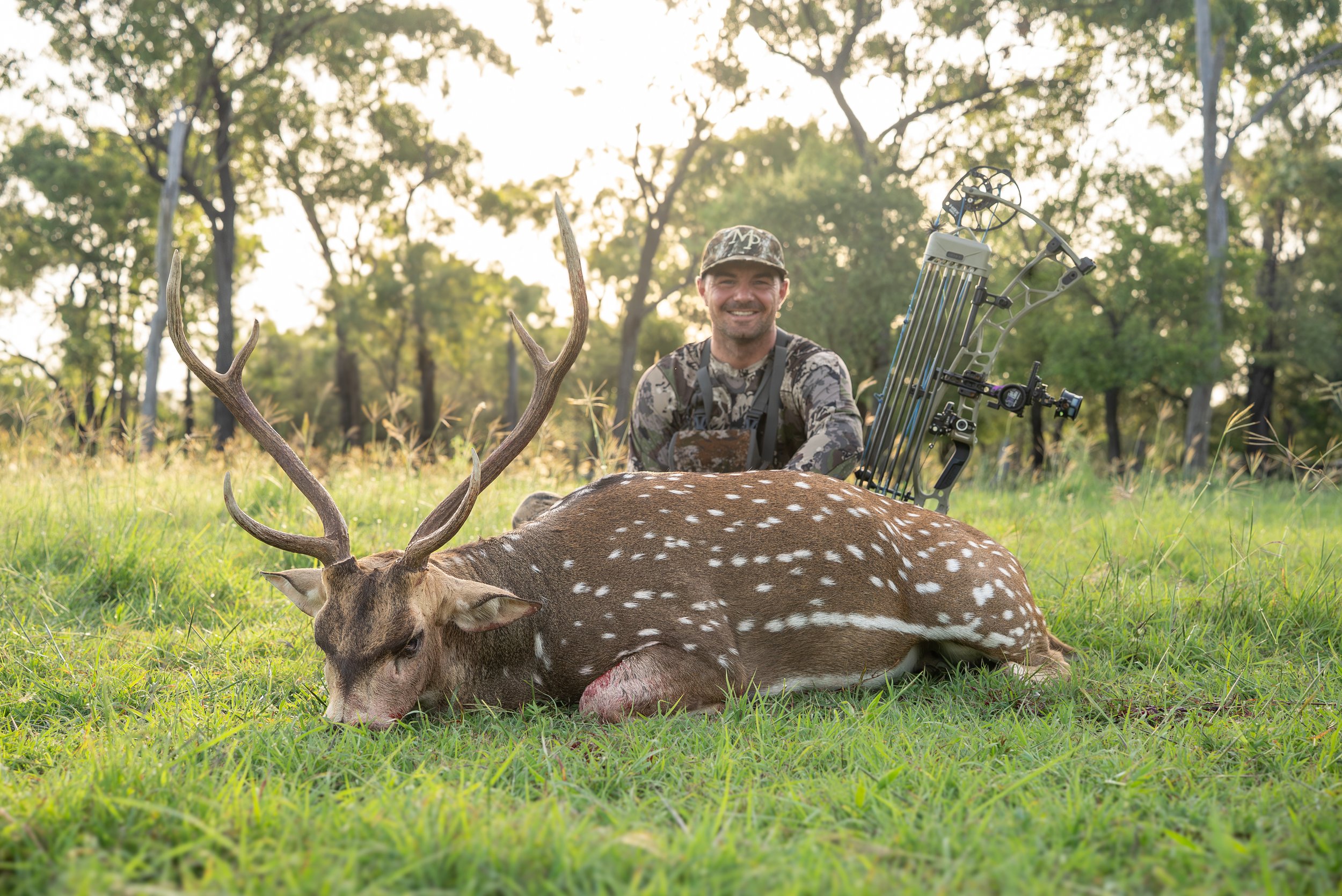 A hunter in camouflage clothing and a cap kneels behind a fallen deer with antlers, smiling, in a forest clearing.