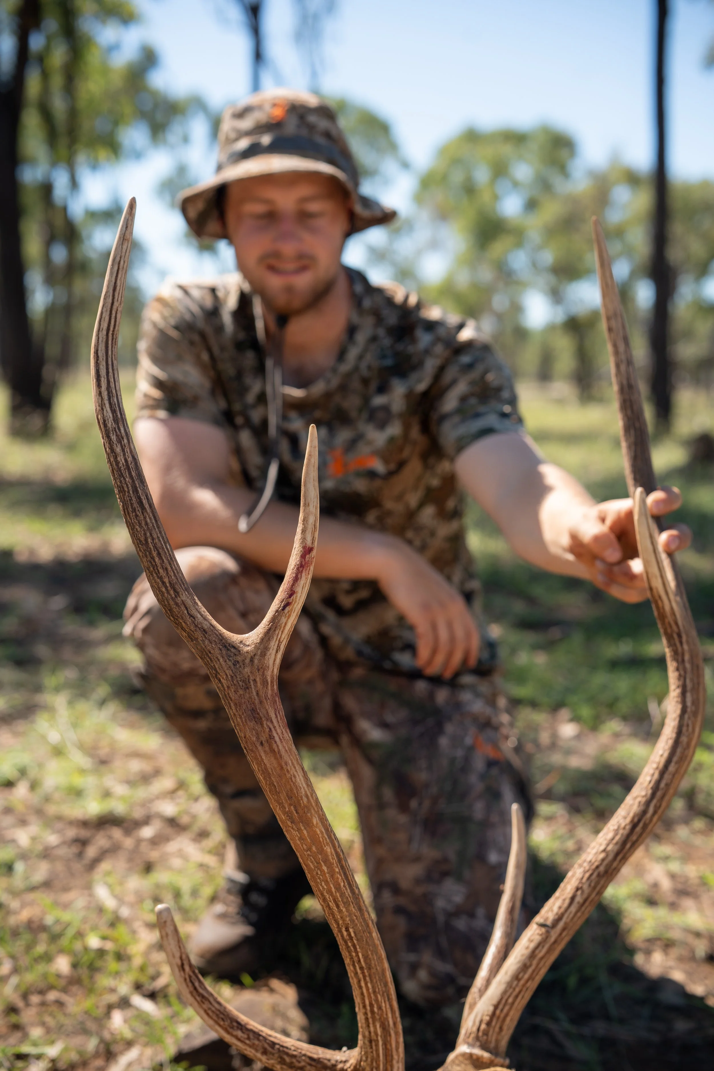 A man in camouflage clothing and a hat kneels on the ground outdoors, holding a large deer antler with multiple points.