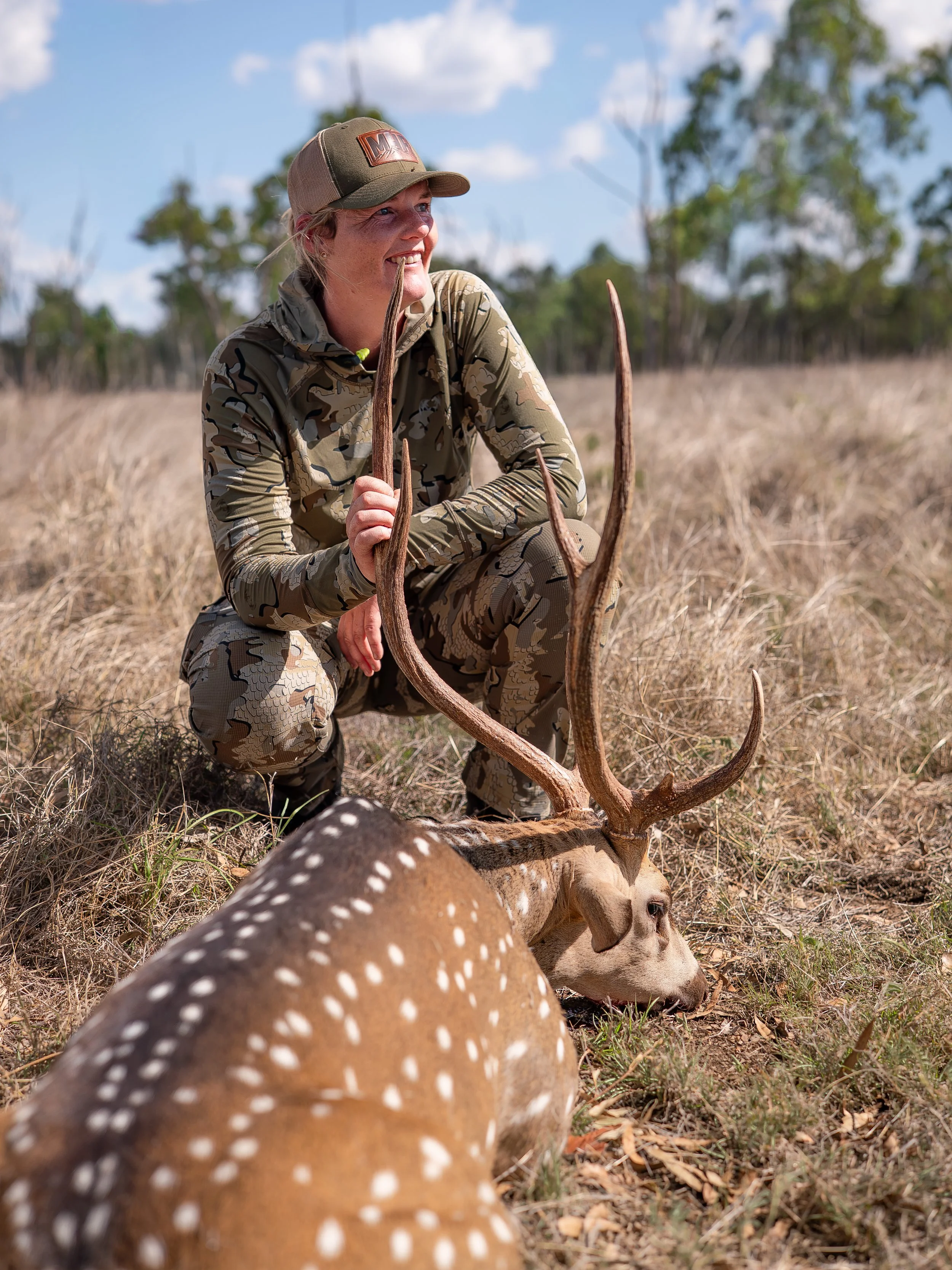A woman dressed in camouflage hunting clothing kneeling in dry grass, smiling, and holding antlers of a recently hunted deer, with the deer lying on the ground. There are trees and a partly cloudy sky in the background.