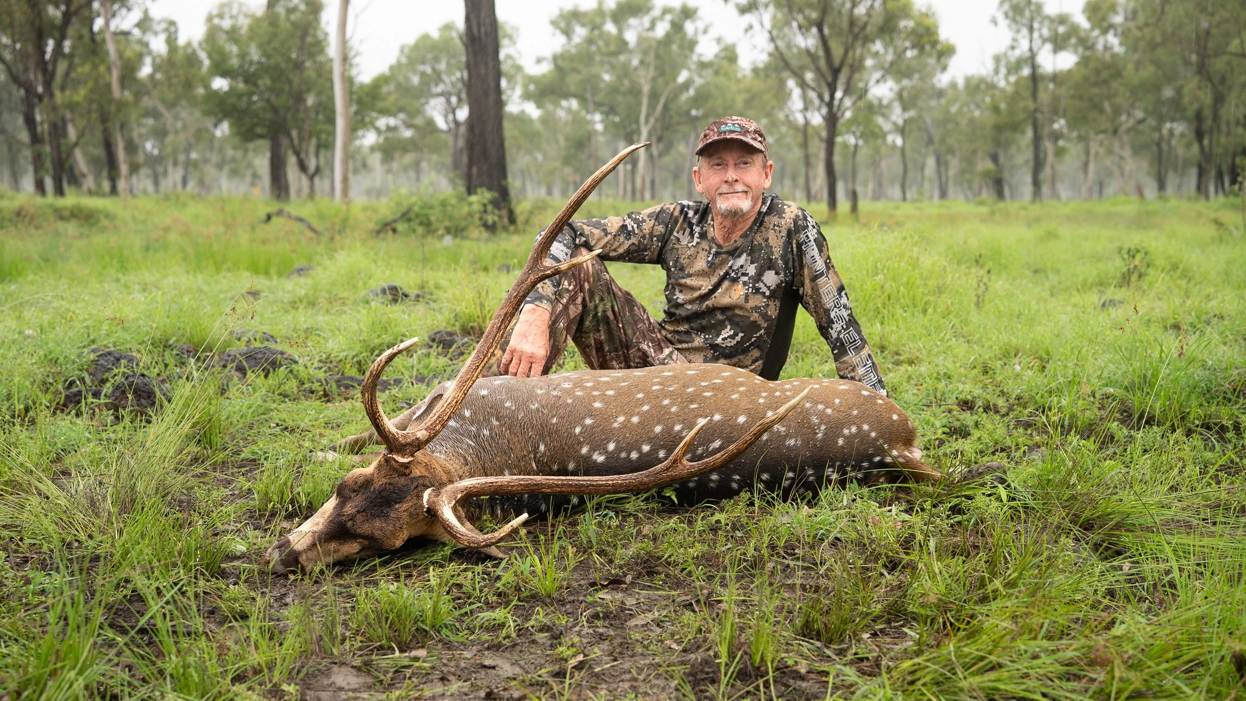 A man dressed in camouflage clothing sitting next to a large dead deer with antlers, in a grassy forested area.