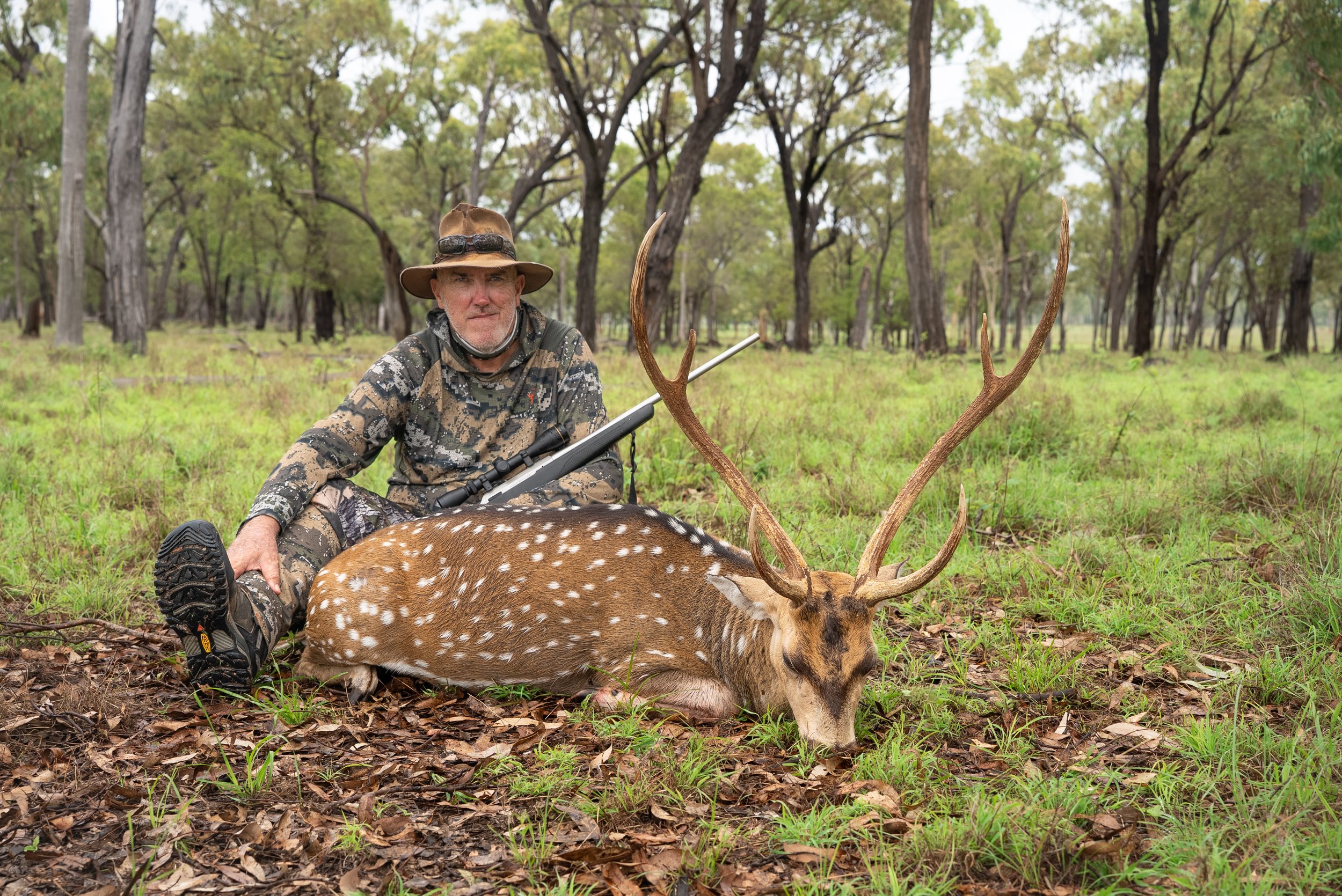 A man dressed in camouflage hunting gear, sitting on the ground in a forest, after hunting a spotted deer.