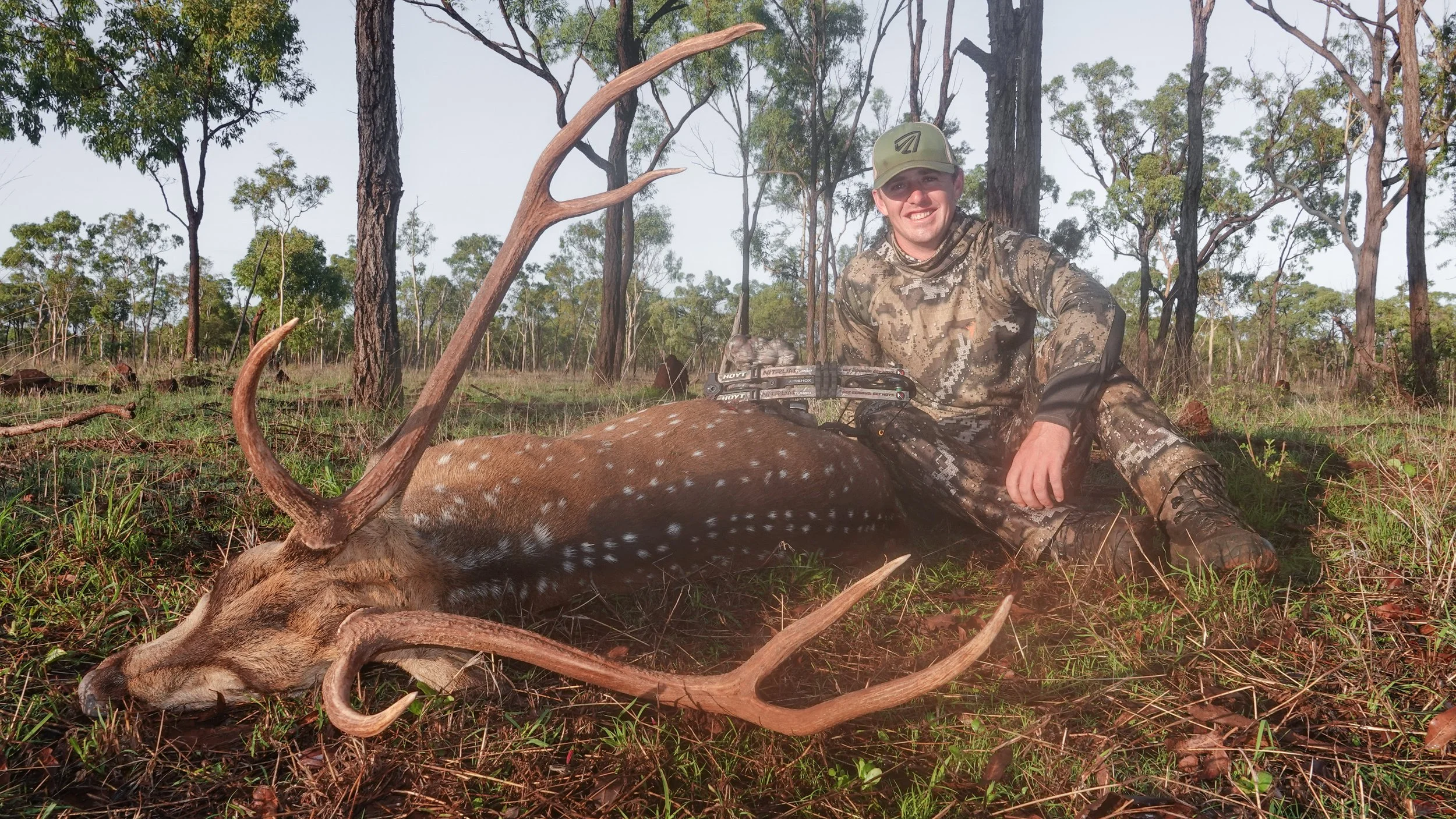 A man in camouflage clothing and a green cap sitting on the ground outdoors in a forest, smiling near a recently hunted deer with antlers lying on the grass.