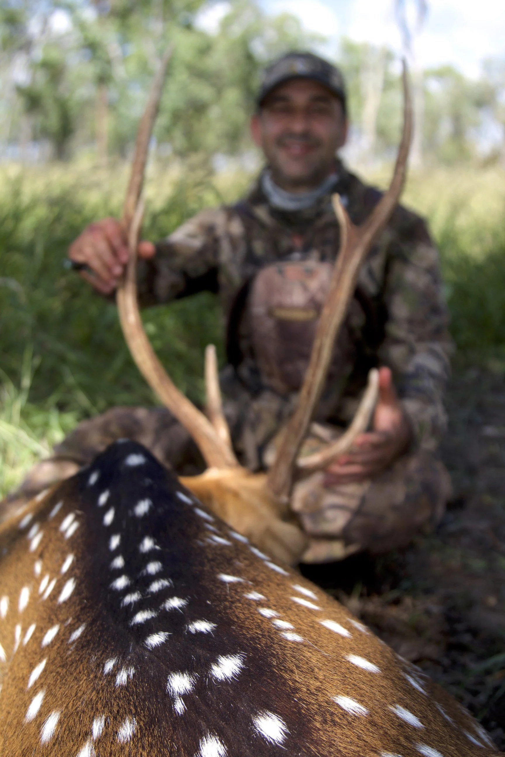 A person in camouflage gear kneeling in a forest, smiling, holding a large set of antlers, with part of a black and white speckled animal in the foreground.