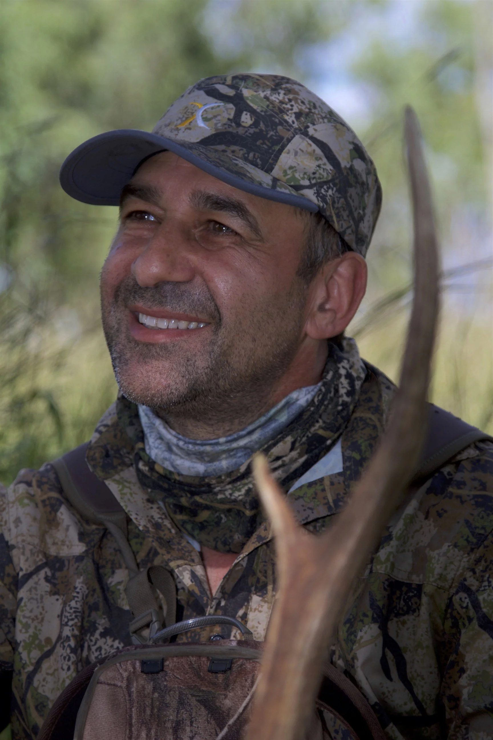 Close-up of a man wearing a camouflage outdoor hunting or hiking gear and a camouflage cap with a moon and star logo, smiling in a natural outdoor setting with blurred greenery in the background.