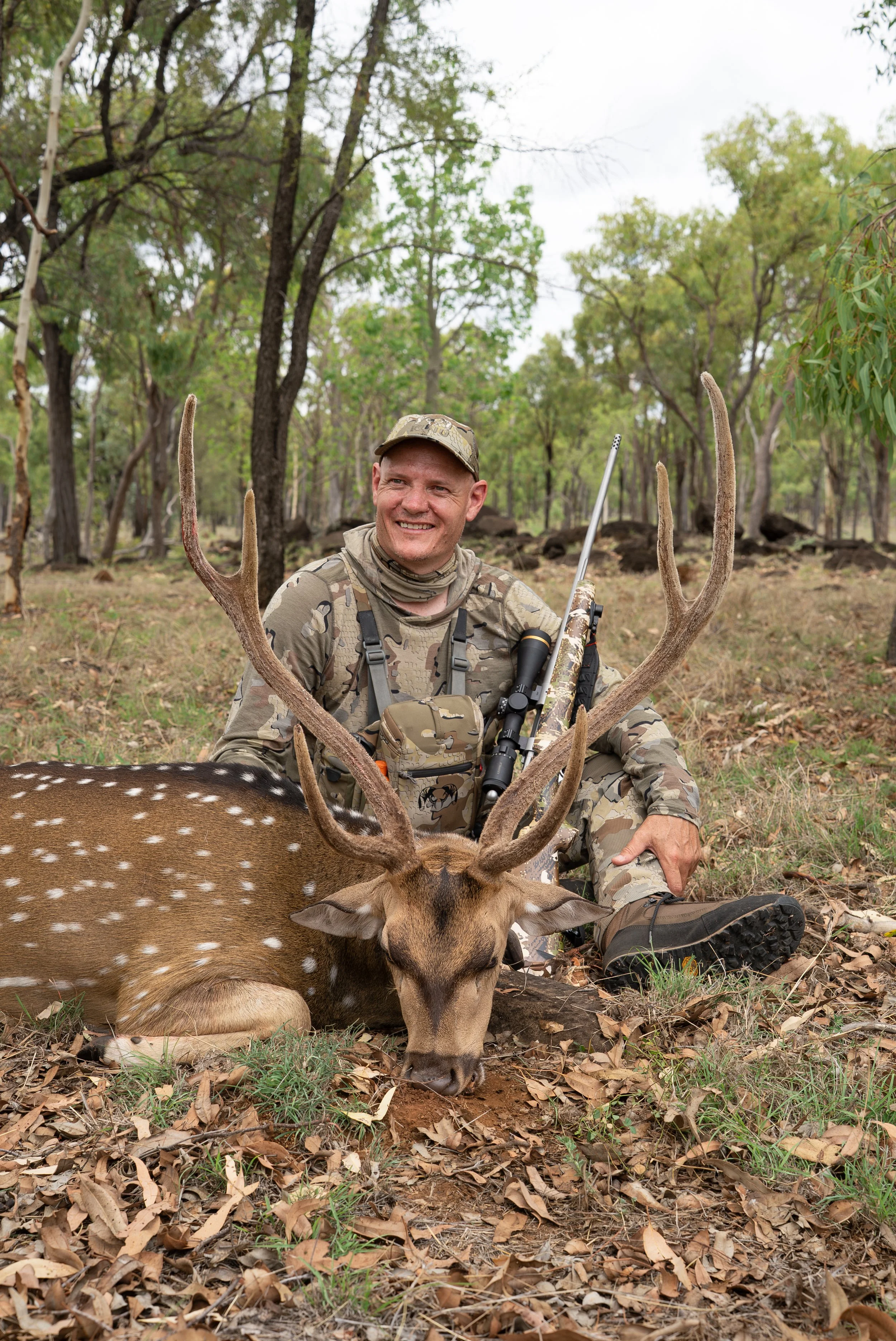 A man dressed in camouflage clothing smiling while sitting on the ground in a forest, holding a large buck with antlers that is lying on the forest floor.