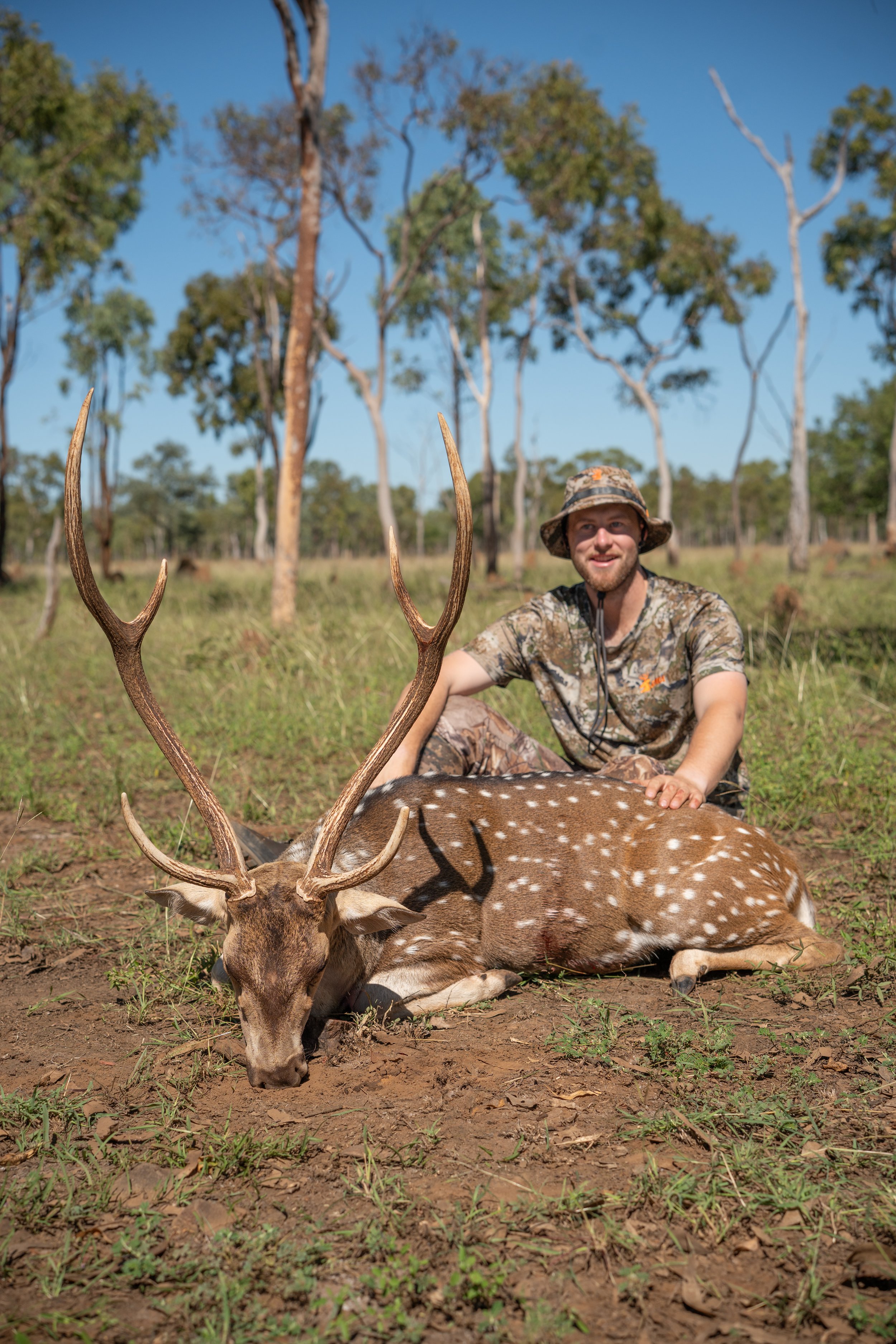 A man wearing camouflage clothing and a camouflage hat is crouching on the ground, smiling, with his hand resting on the back of a dead spotted deer. The deer is lying on the ground with large antlers visible, and its head is resting on the earth. Th