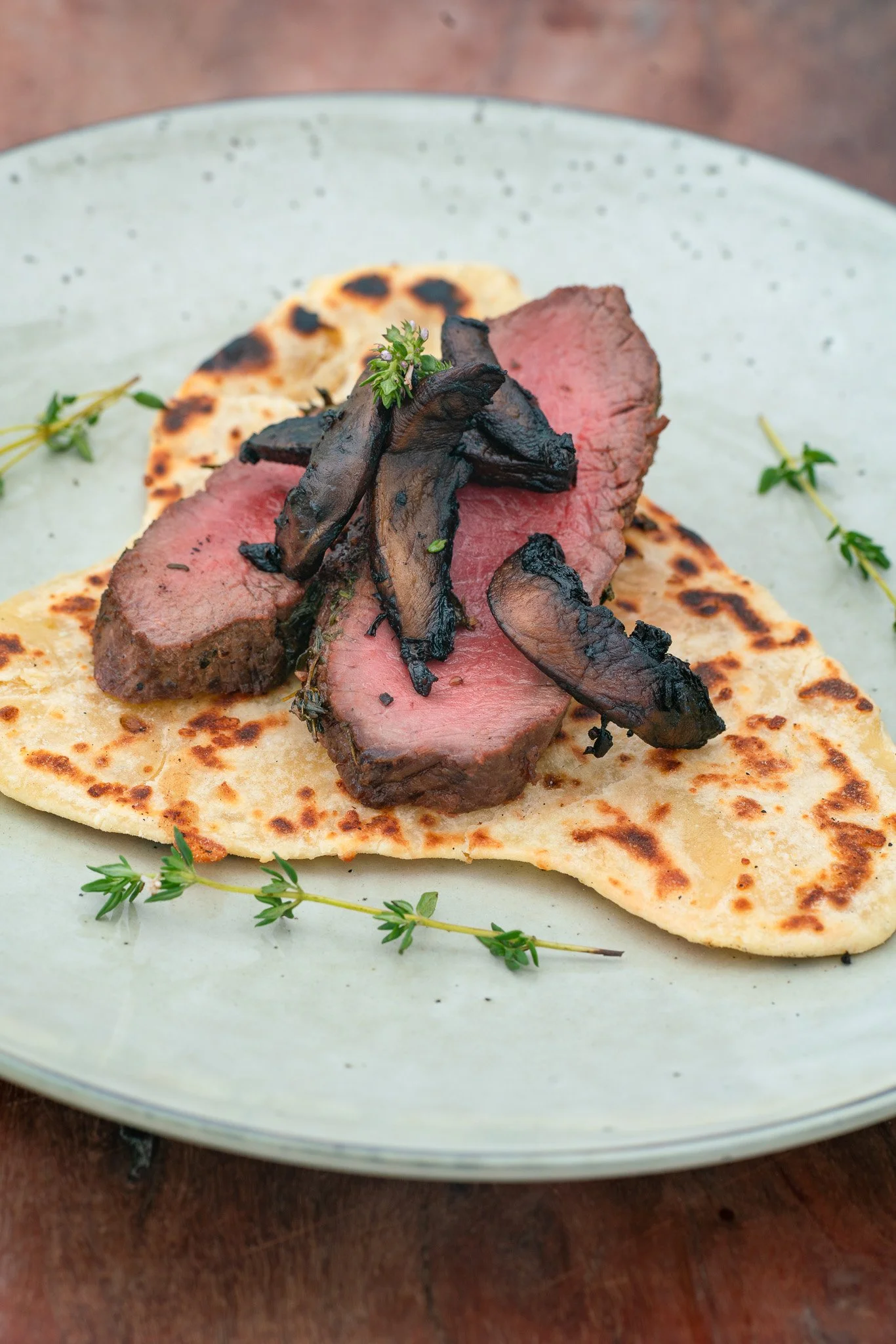 Close-up of a piece of flatbread topped with grilled steak slices, sautéed mushrooms, and garnished with fresh herbs, served on a light green plate.
