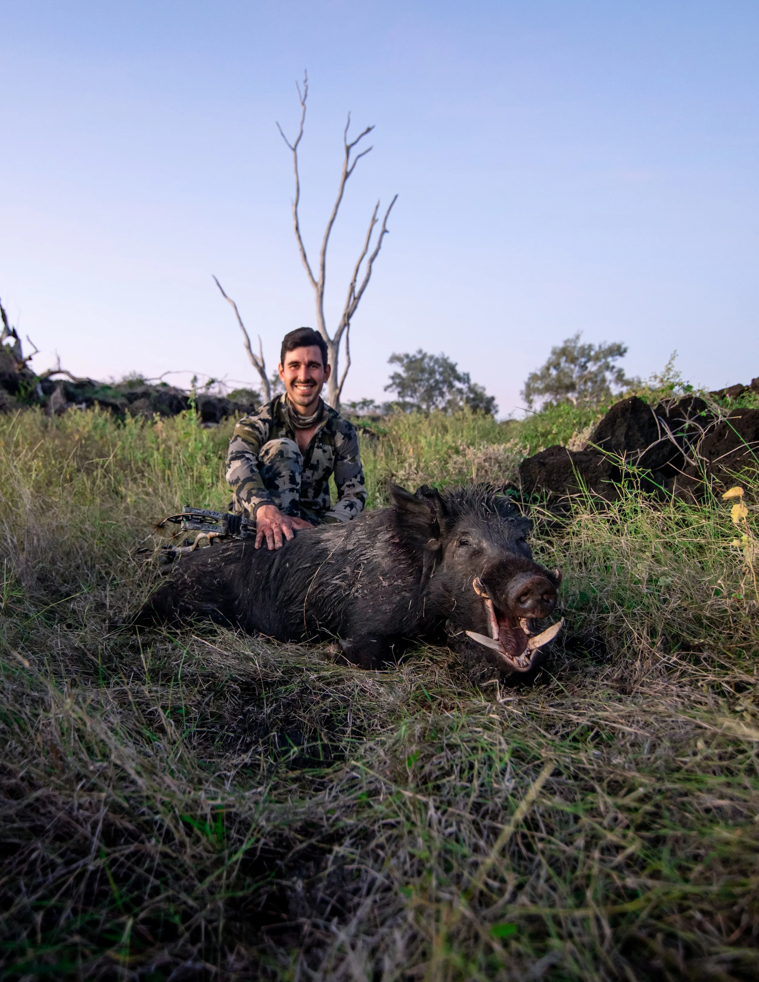 Smiling man in camouflage clothing sitting next to a dead wild boar in a grassy outdoor area with leafless trees in the background.