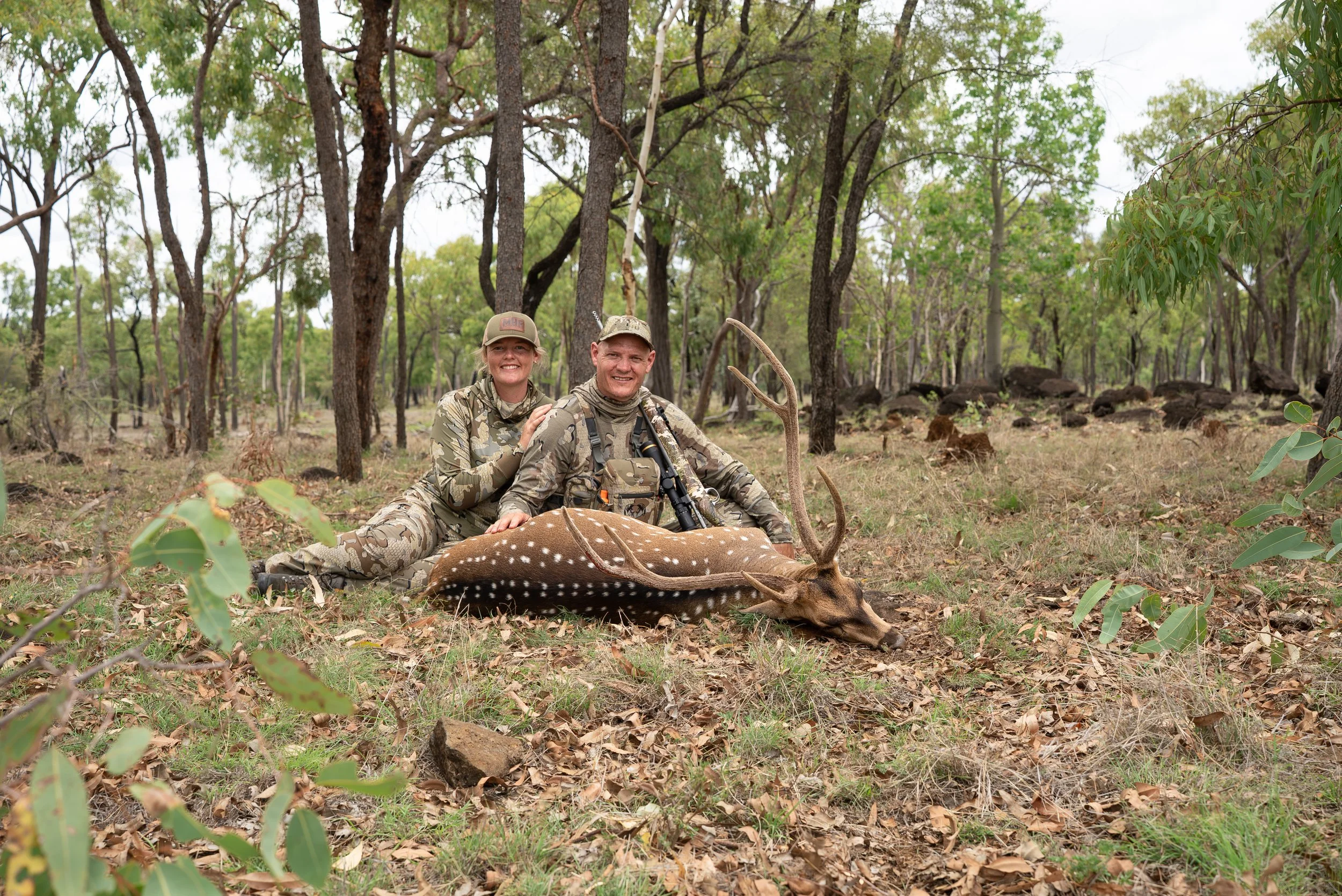 Two hunters, a woman and a man, sitting in a forest clearing, smiling at the camera with a dead spotted deer lying in front of them. They are dressed in camouflage hunting gear and hats, surrounded by trees and rocks.