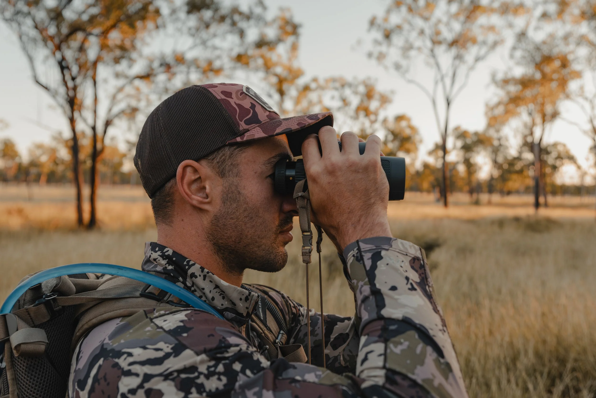 A man wearing camouflage outdoor clothes and a camouflage cap, seen holding binoculars up to his eyes in an autumn outdoor setting.