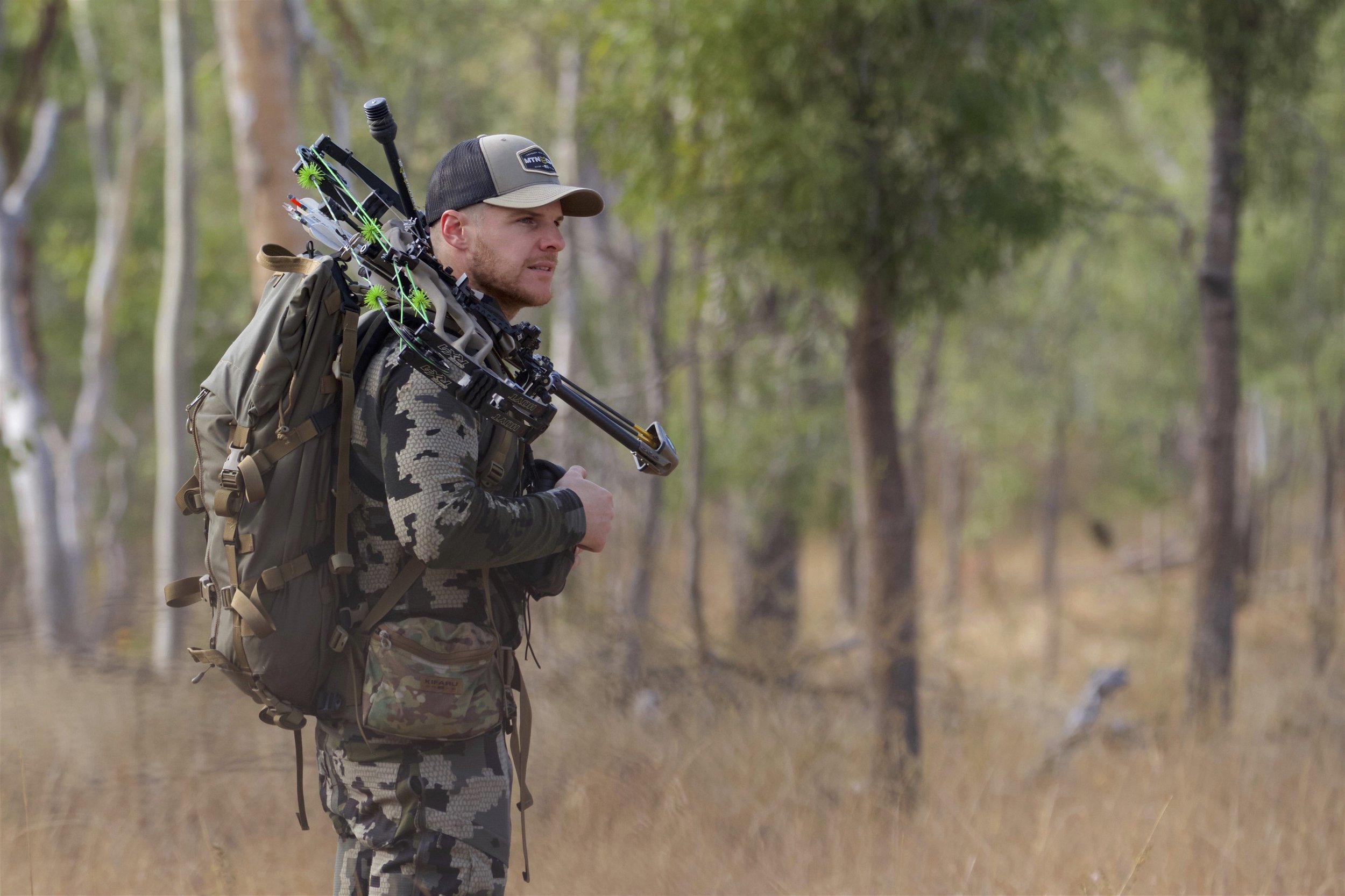 A man dressed in camouflage clothing and a baseball cap walks through a wooded area with a large backpack carrying a compound bow, aiming to go hunting.