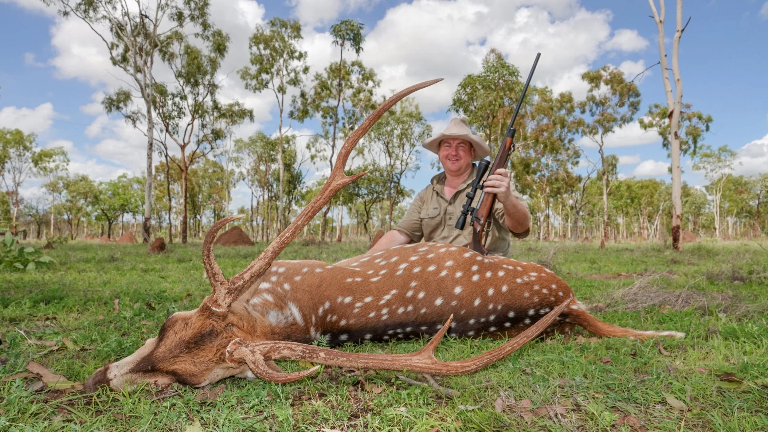 A man in safari attire sitting in a forest clearing, smiling, holding a rifle, next to a fallen spotted deer with large antlers.