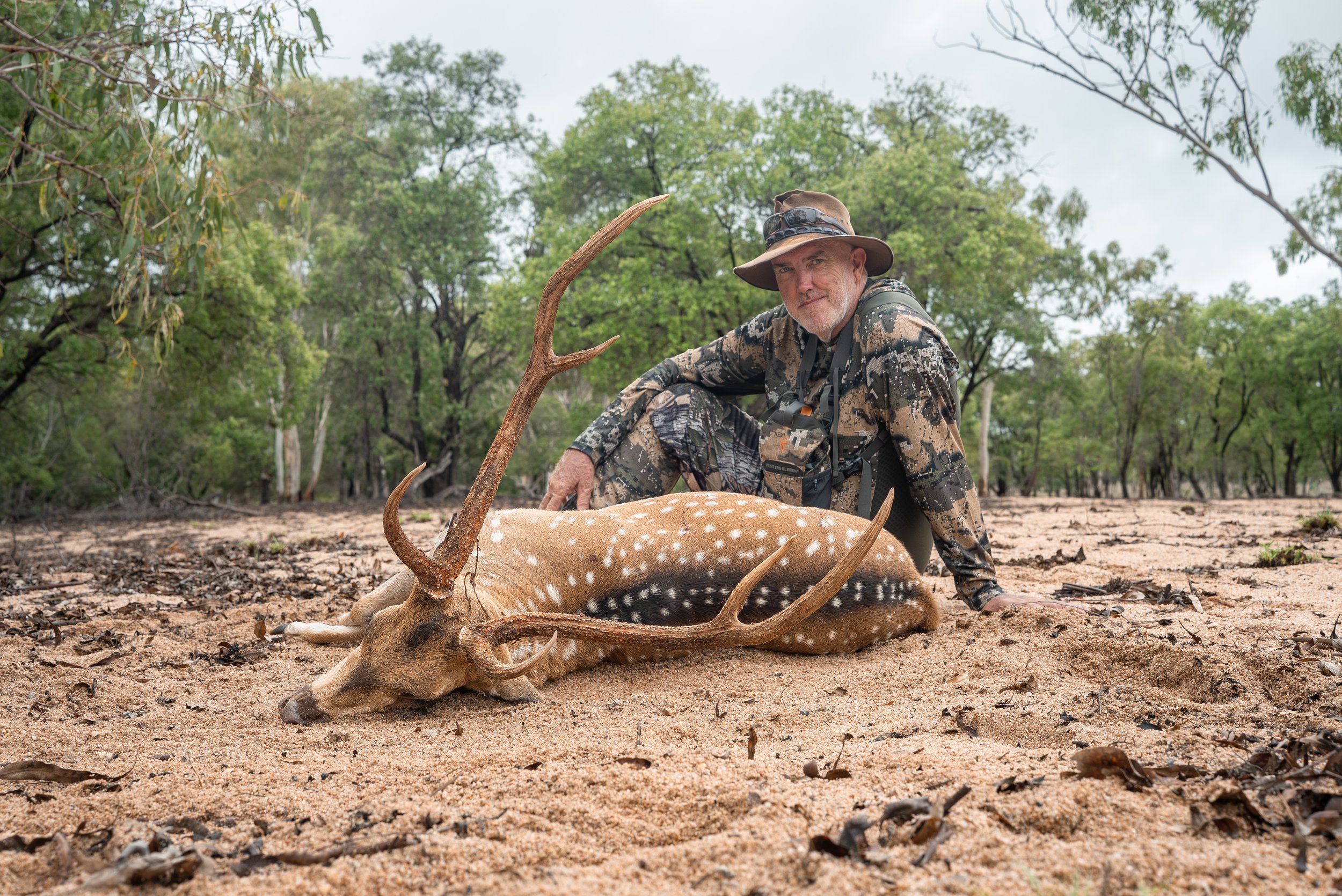 A man dressed in camouflage clothing and a wide-brimmed hat kneels beside a freshly hunted deer with large antlers, lying on dry, sandy ground surrounded by trees and bushes.