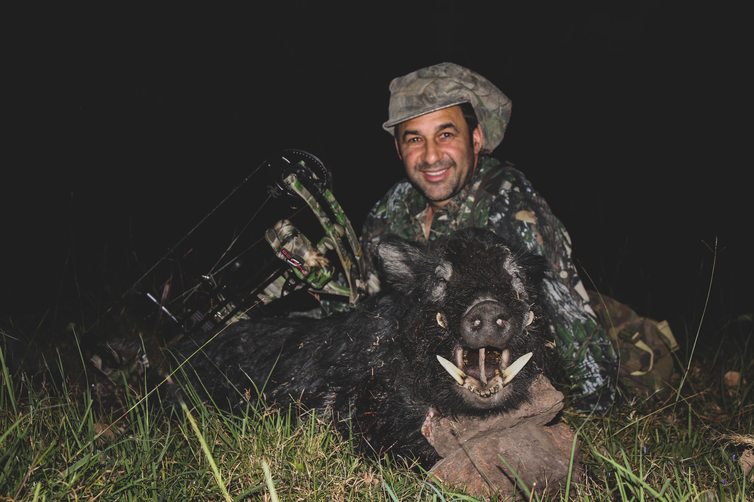 A man dressed in camouflage hunting gear and a hat, smiling while kneeling in grass during nighttime, holding a large wild boar with sharp teeth and tusks, which appears to be dead.