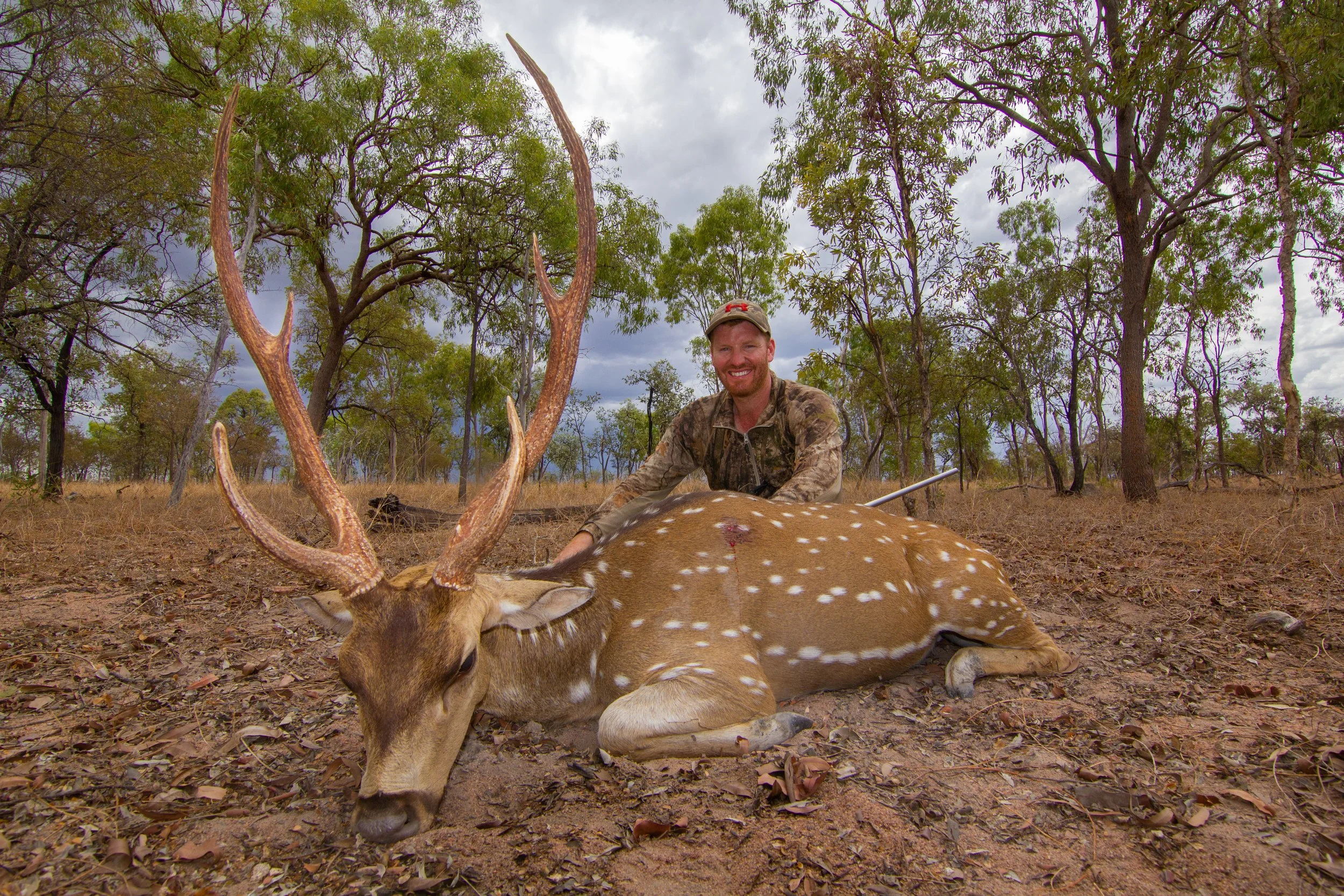 A man in camouflage clothing and a cap is smiling while kneeling behind a fallen deer with antlers in a wooded area during the daytime.