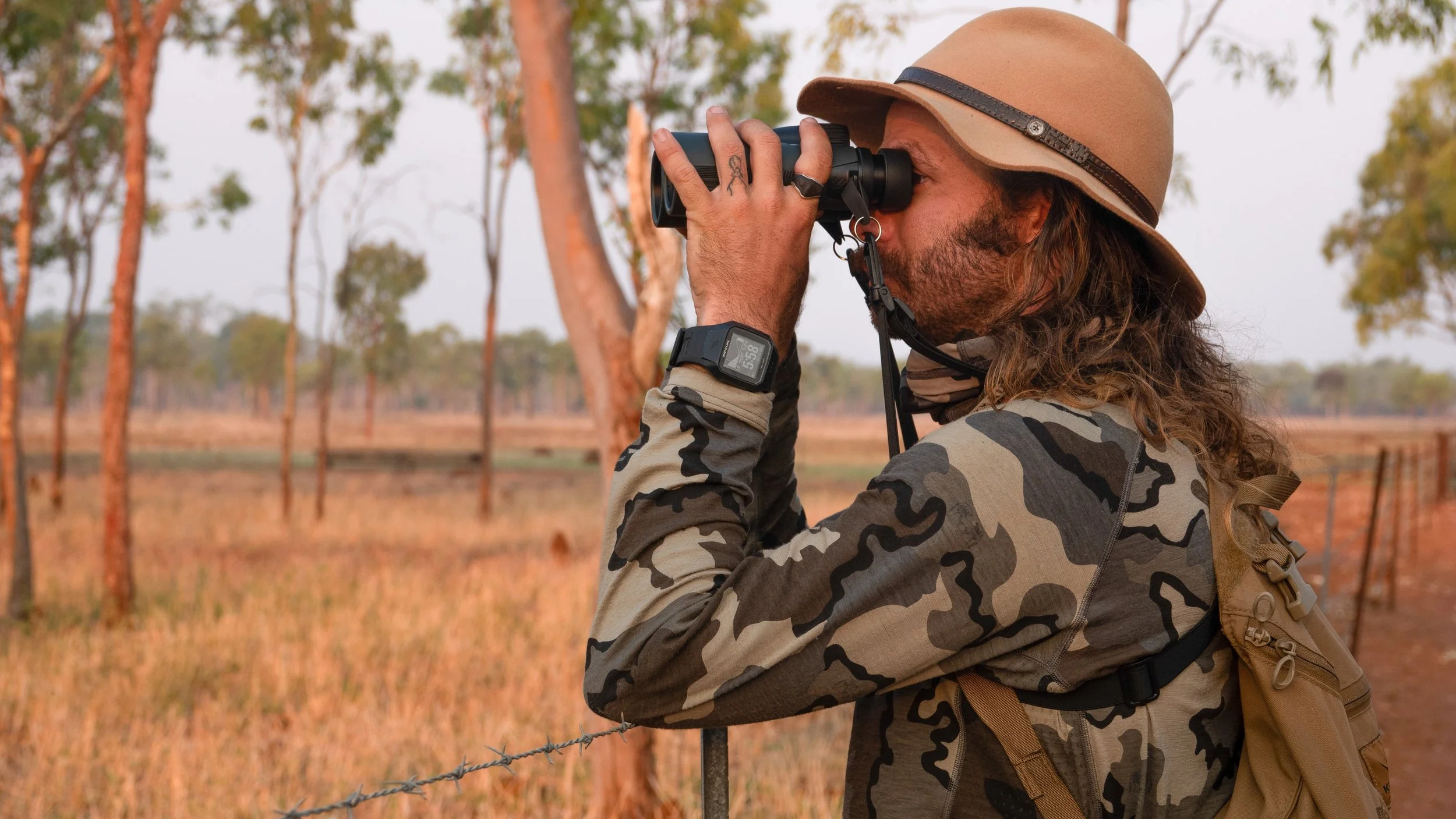 Person with long hair, wearing a tan hat and camouflage shirt, looking through binoculars outdoors in a rural area with trees and a fence.
