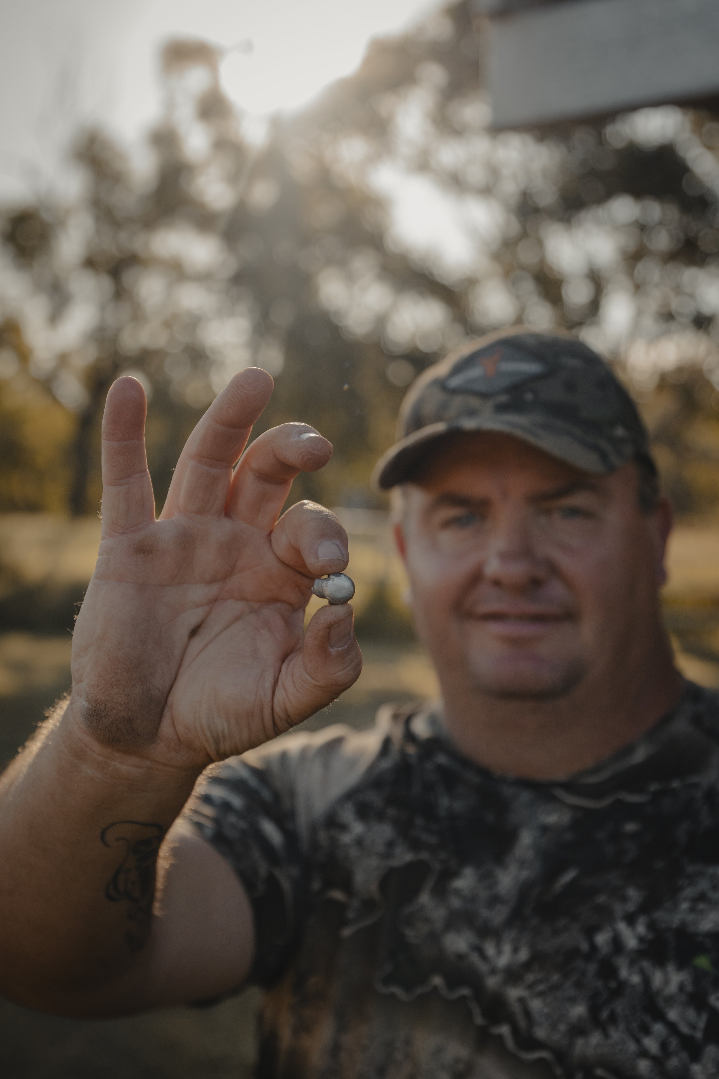 A man with a camouflage baseball cap and a tattoo on his arm holds a small silver fish toward the camera. The background is blurred with trees and warm sunlight.