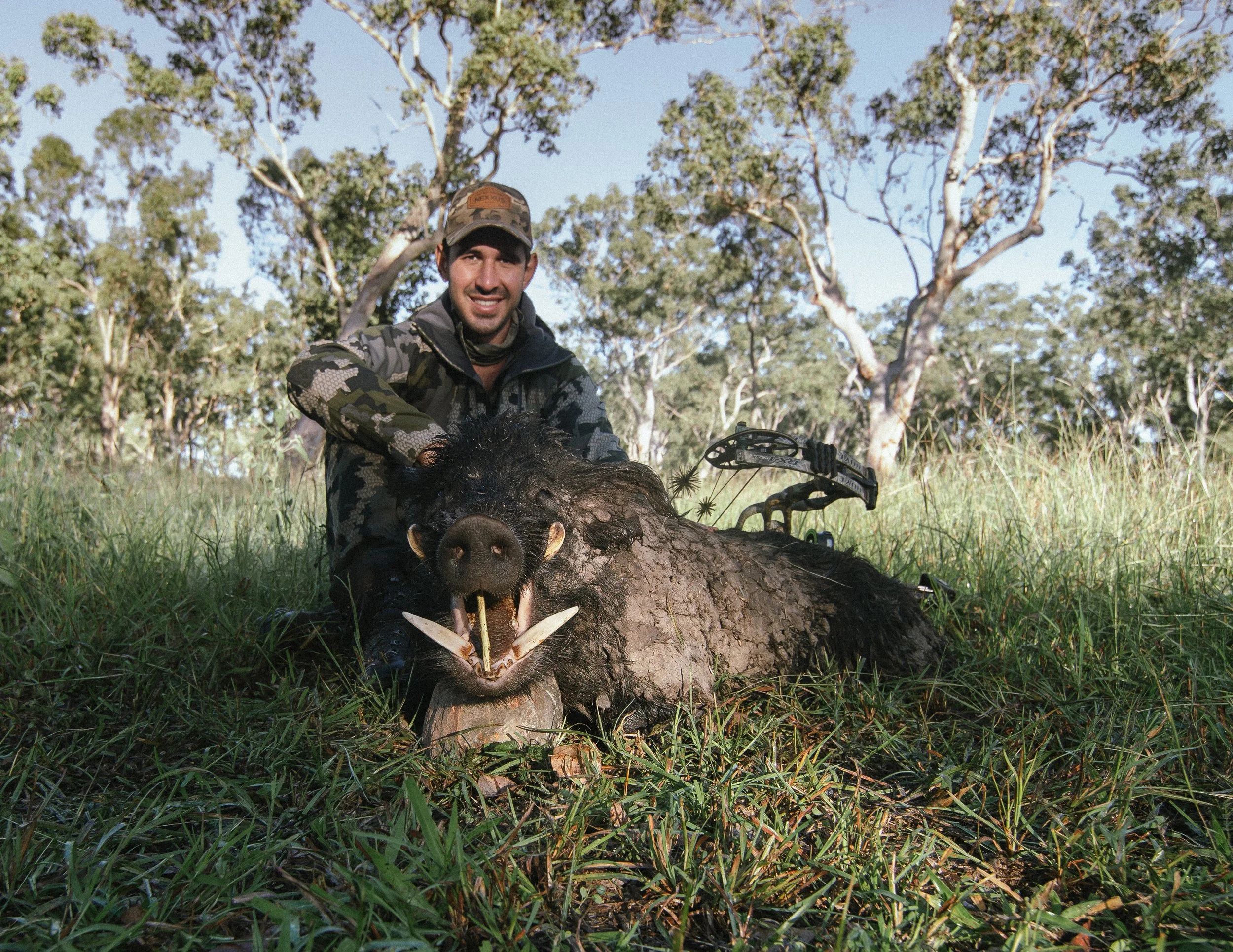 A man wearing camouflage clothing and a cap sitting on the ground outdoors next to a fallen tree trunk with a bow and arrow, with trees and blue sky in the background.