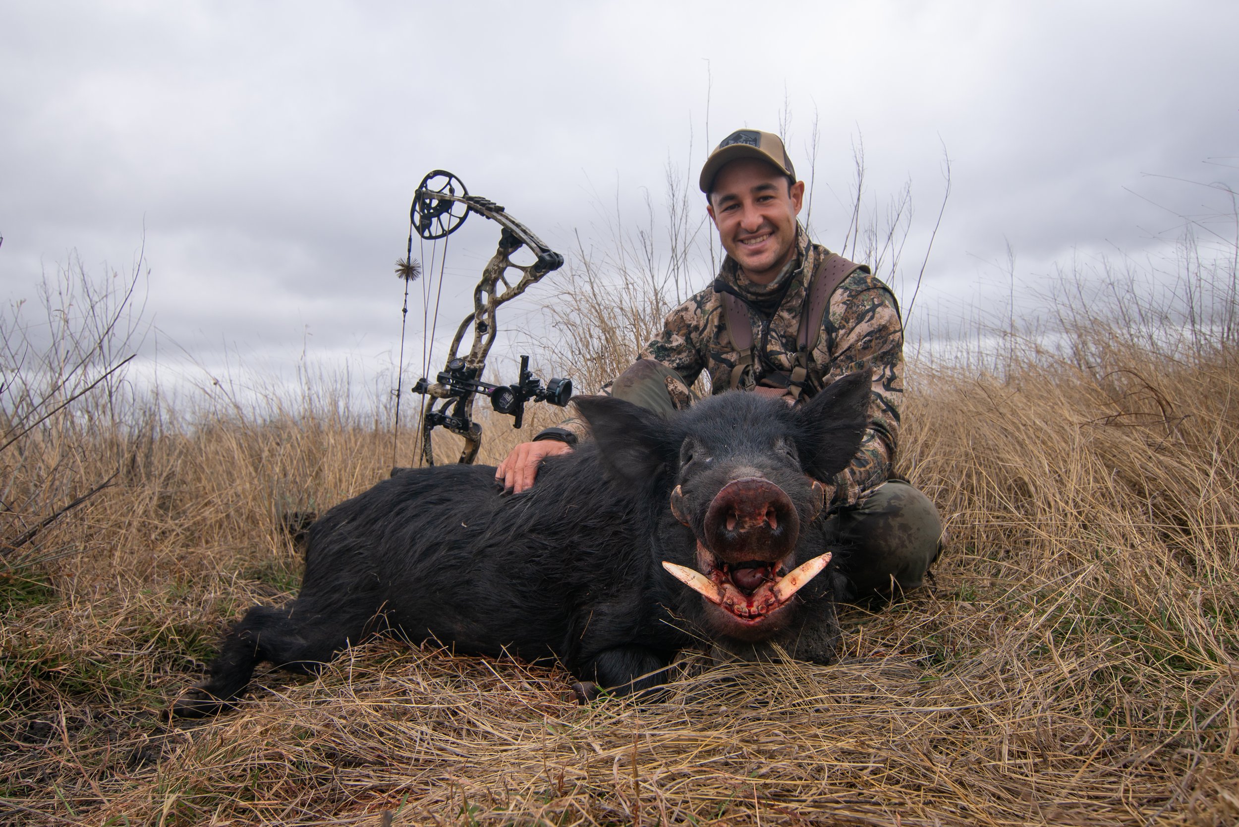 A smiling man in camouflage clothing and a cap sitting outdoors in tall grass, holding a dead pig with a bow and arrow nearby.