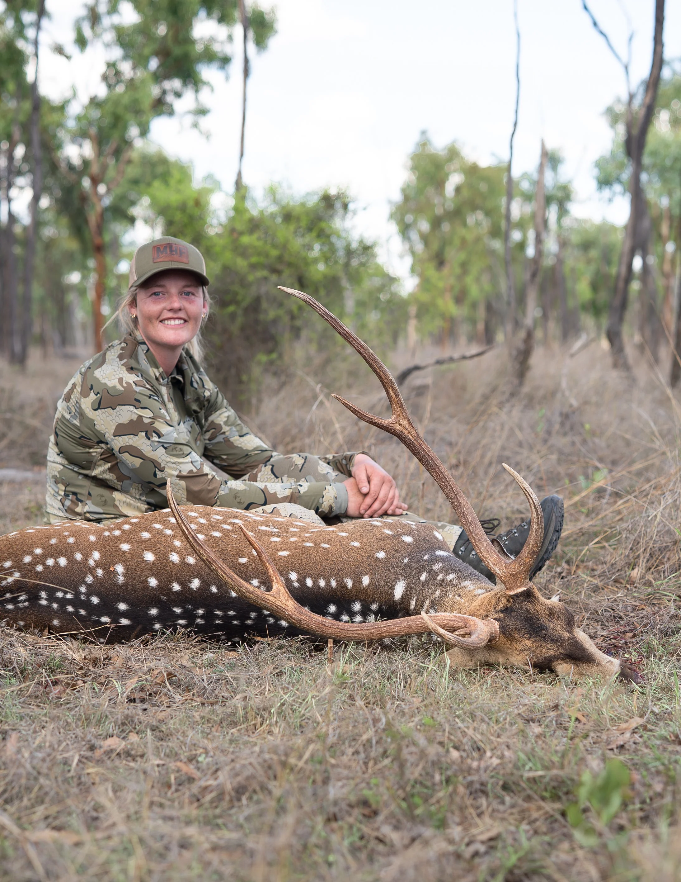 A woman dressed in camouflage sitting on the ground in a wooded area, smiling, with a large deer with antlers lying on the ground beside her.