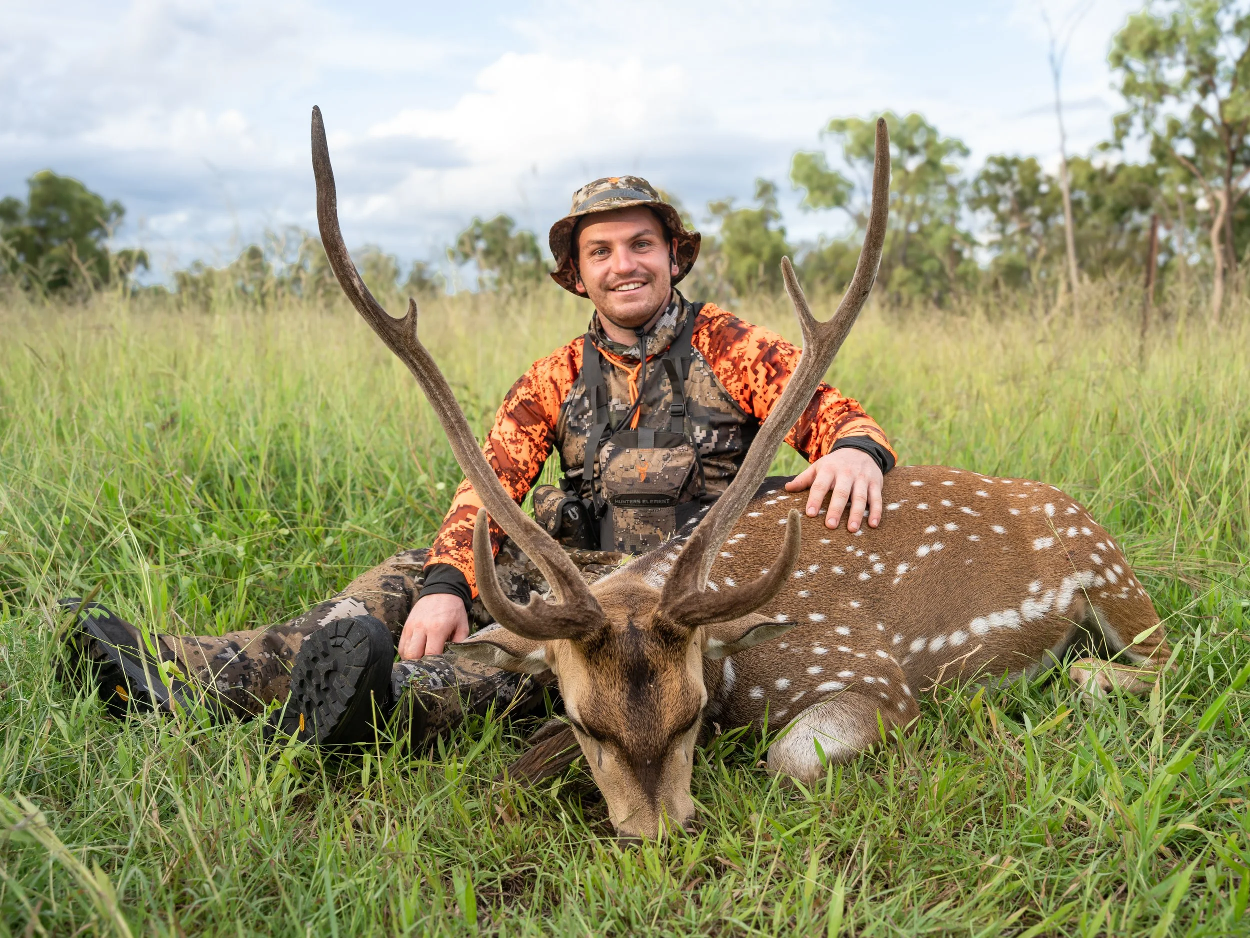 A man dressed in camouflage hunting gear sitting in tall grass with a deer, holding its antlers, outdoors in a field with trees and cloudy sky in the background.