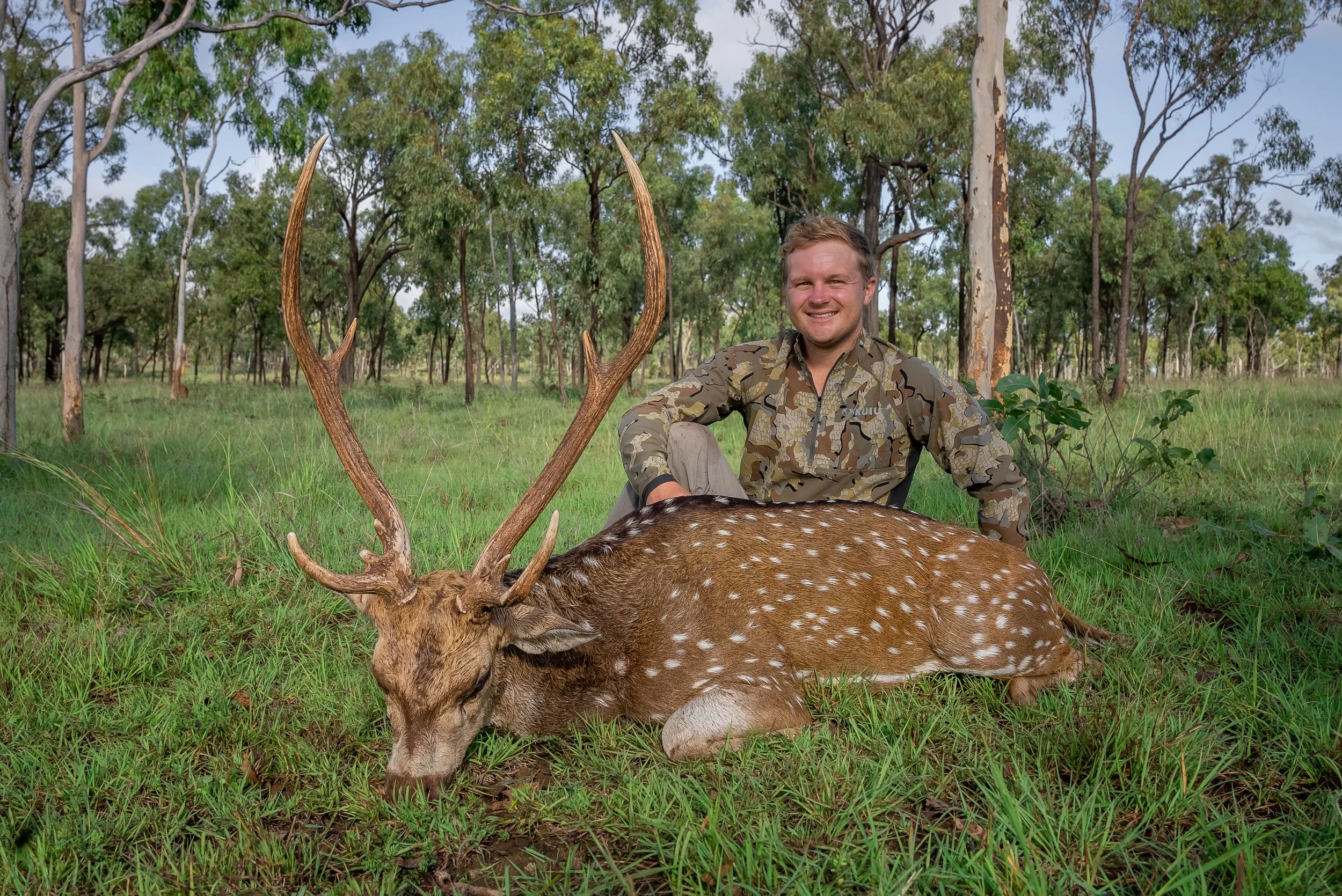 A man in camouflage clothing sitting in a forested area with a large deer that has prominent antlers, lying on the grass in front of him.