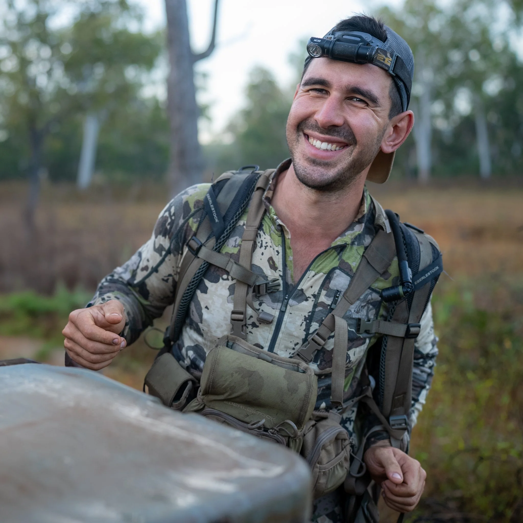 A man smiling outdoors wearing a headlamp, camouflage shirt, and backpack, extending his hand toward the camera in a wooded area.