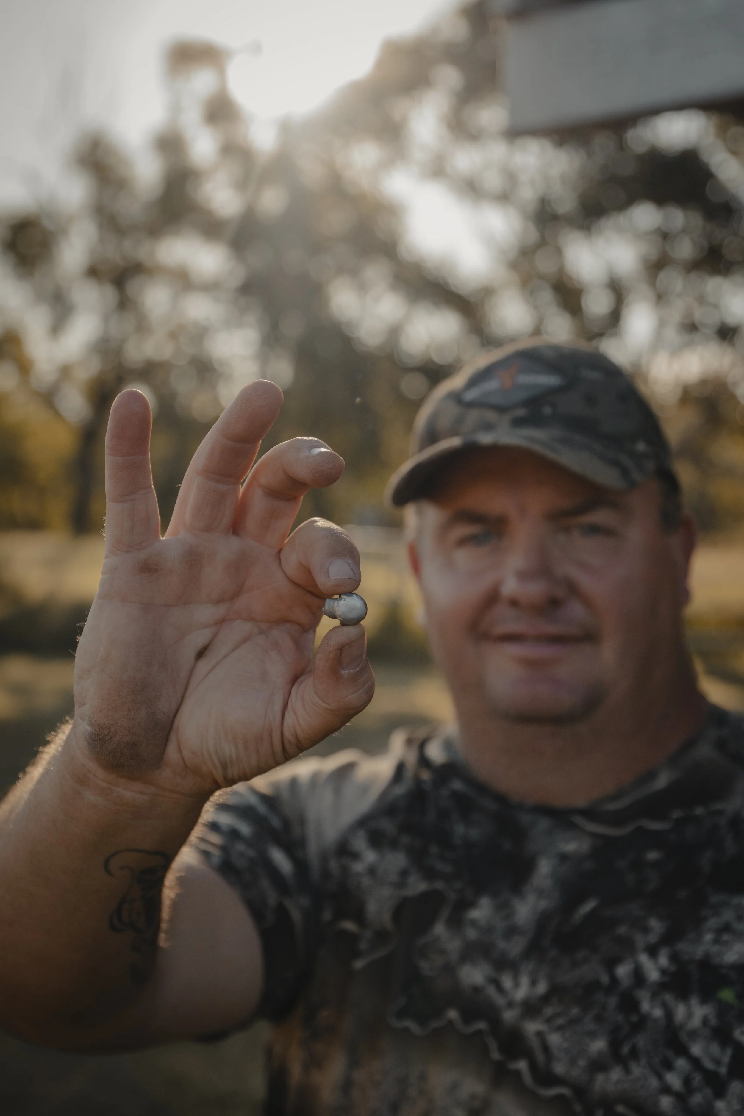 A man wearing a camouflage baseball cap is holding a shiny silver object between his thumb and index finger, with a blurred background of trees and sunlight.