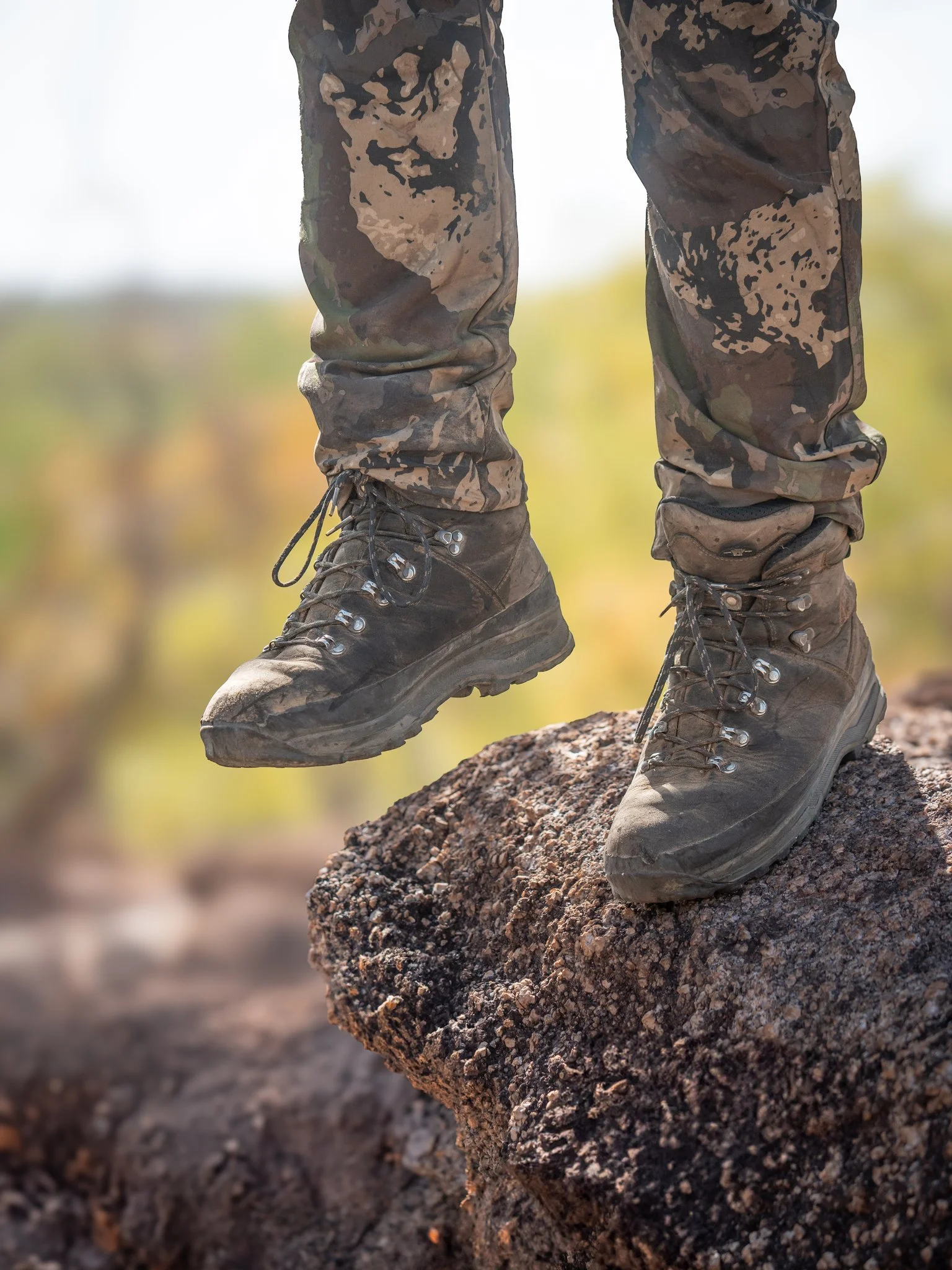 Close-up of muddy hiking boots on a rock amidst an outdoor setting with a blurred background.
