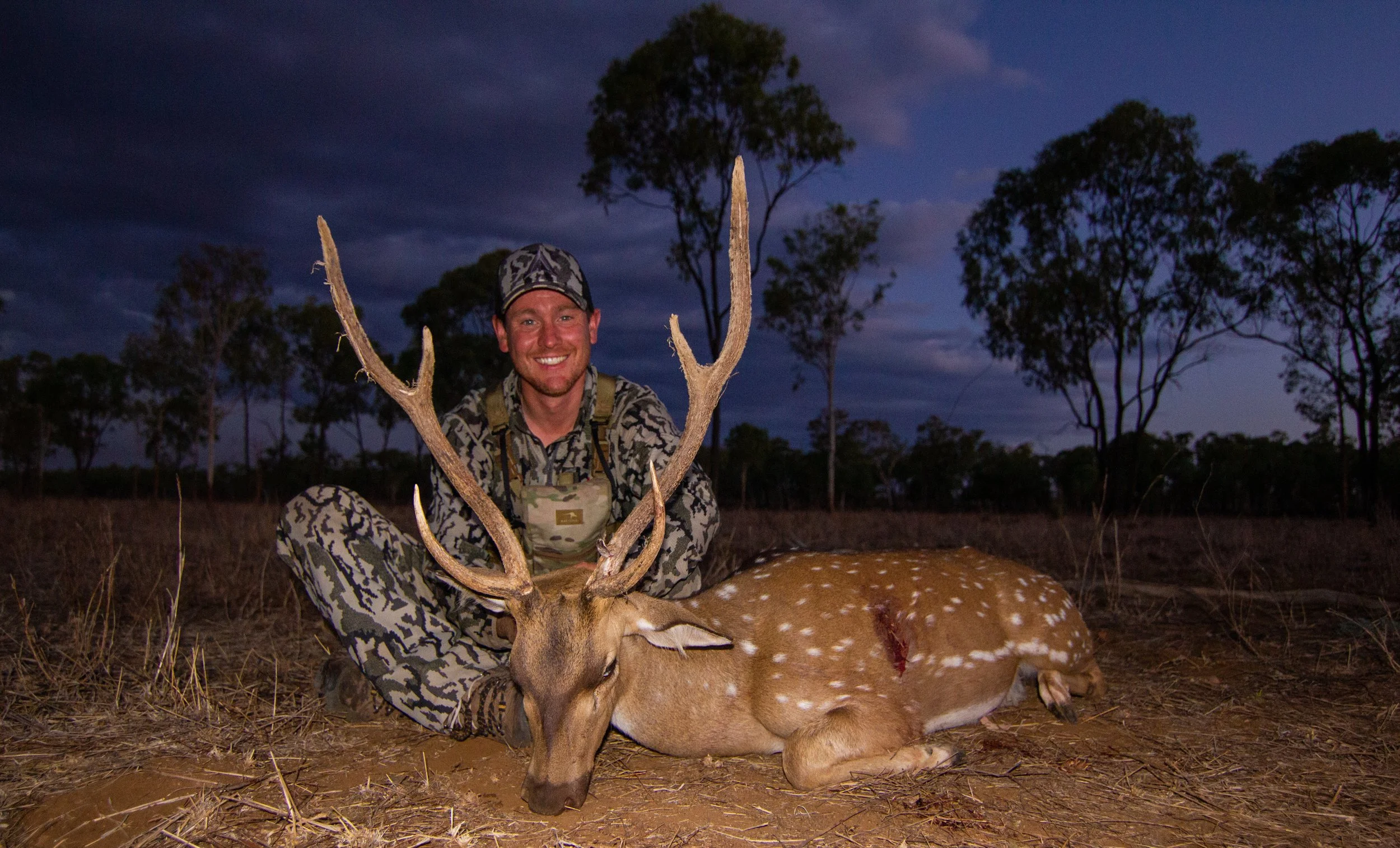 A man in camouflage clothing and a cap kneels on the ground, smiling, beside a fallen deer with antlers, in a wooded outdoor setting at dusk.