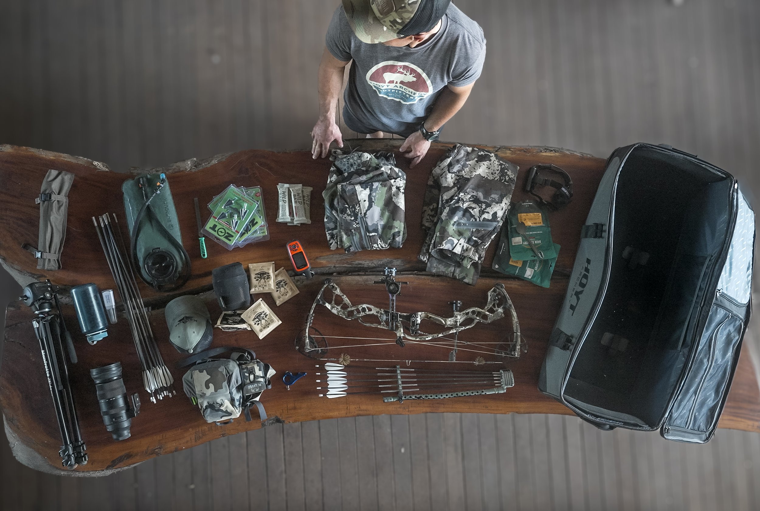 Top-down view of a wooden table with archery equipment and accessories, including a compound bow, arrows, camouflage clothing, a scope, and a carrying case, with a person wearing a T-shirt and camouflage hat standing at the top of the table.
