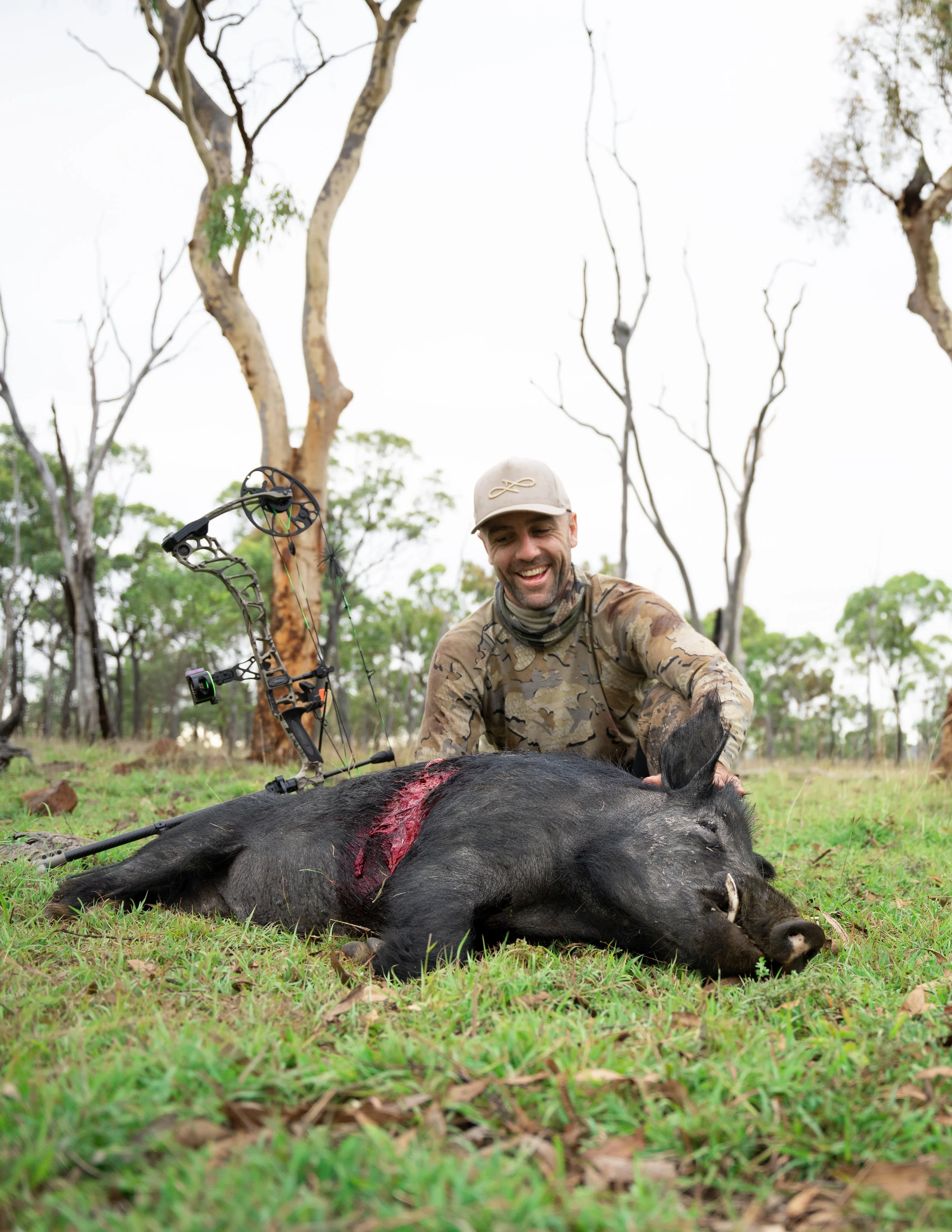 A man dressed in camouflage clothing, smiling, kneeling beside a dead wild boar with visible blood, holding its body in an outdoor wooded area.
