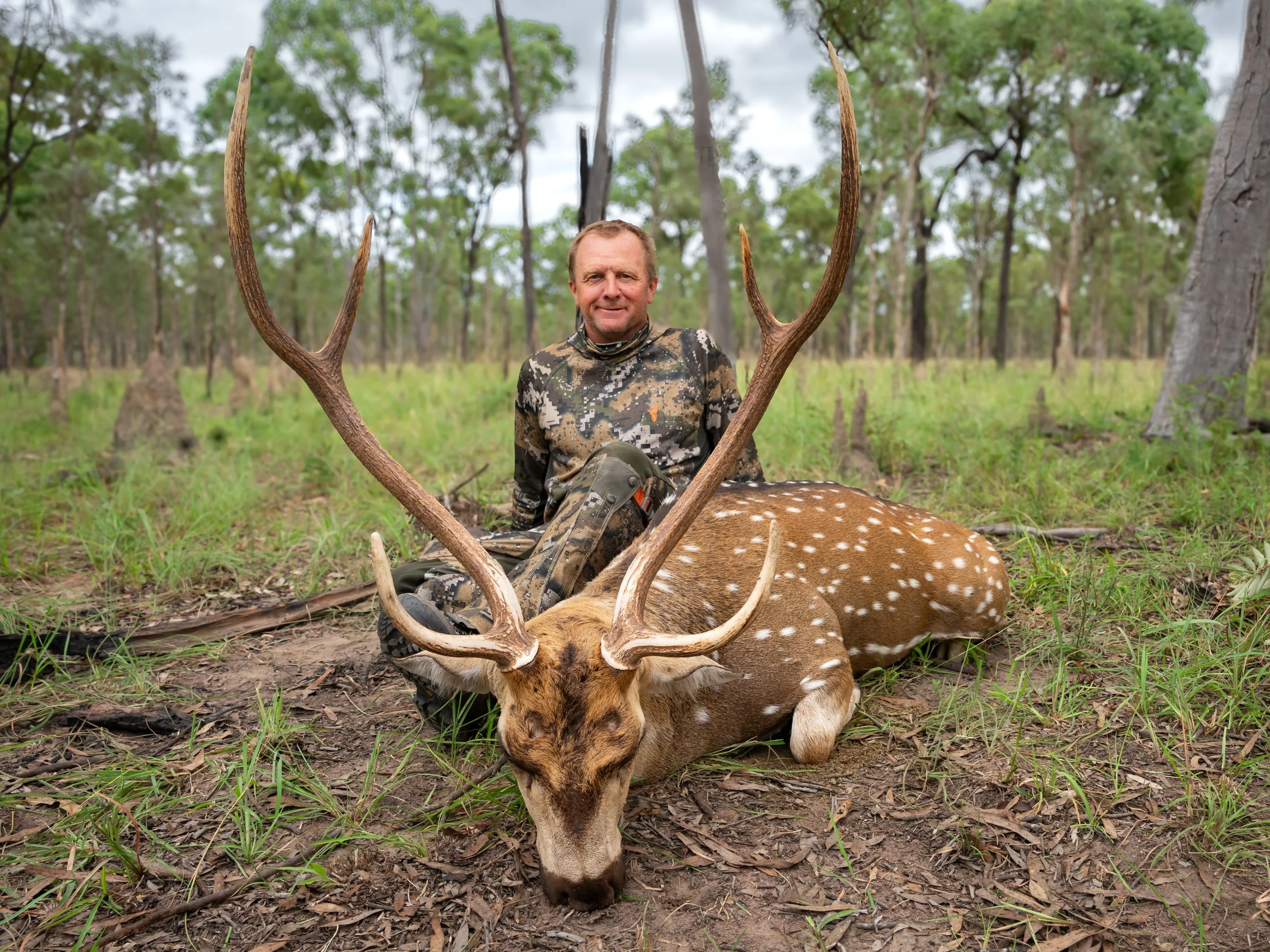 A man dressed in camouflage clothing is sitting on the ground in a wooded area, posing with a large deer with antlers that is lying down in front of him.