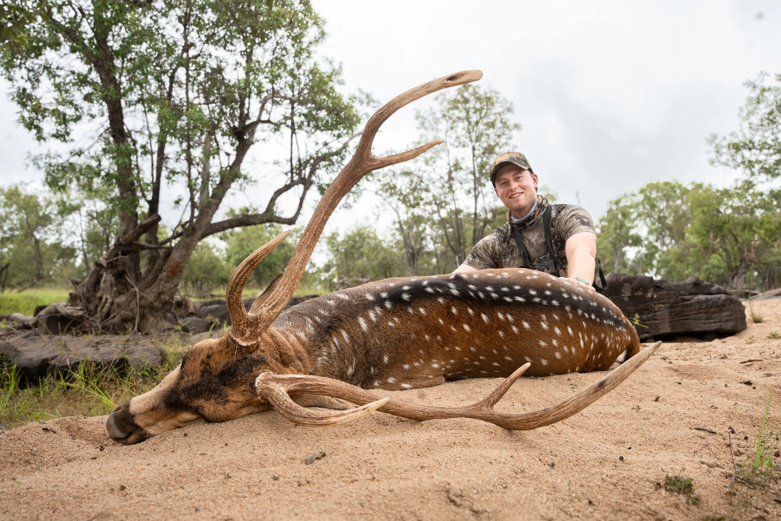 A man in camouflage clothing and hat smiling while kneeling behind a dead deer with large antlers lying on sandy ground in a wooded area.