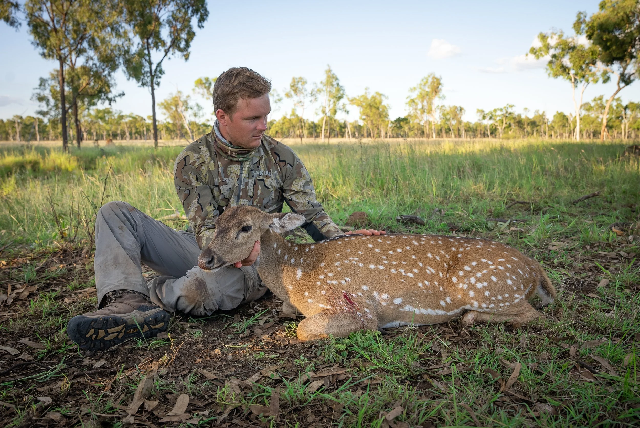 A man in camouflage clothing sitting on the ground in a grassy field holding the head of a dead deer with white spots. The deer has a small wound and appears to have been recently killed.
