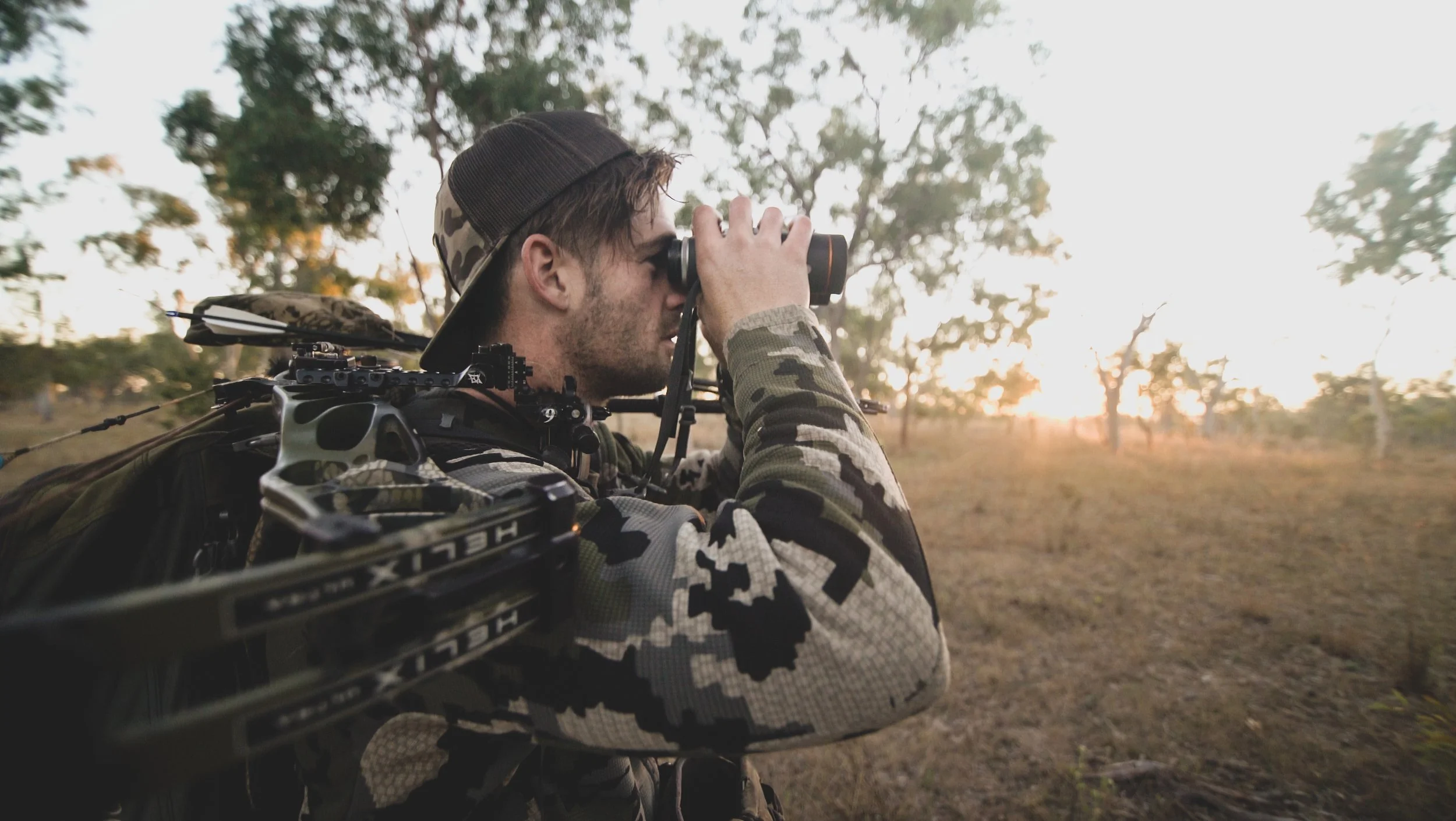 A man in camouflage clothing and a cap looking through binoculars in an outdoor setting during sunset, with trees and a dirt ground in the background.