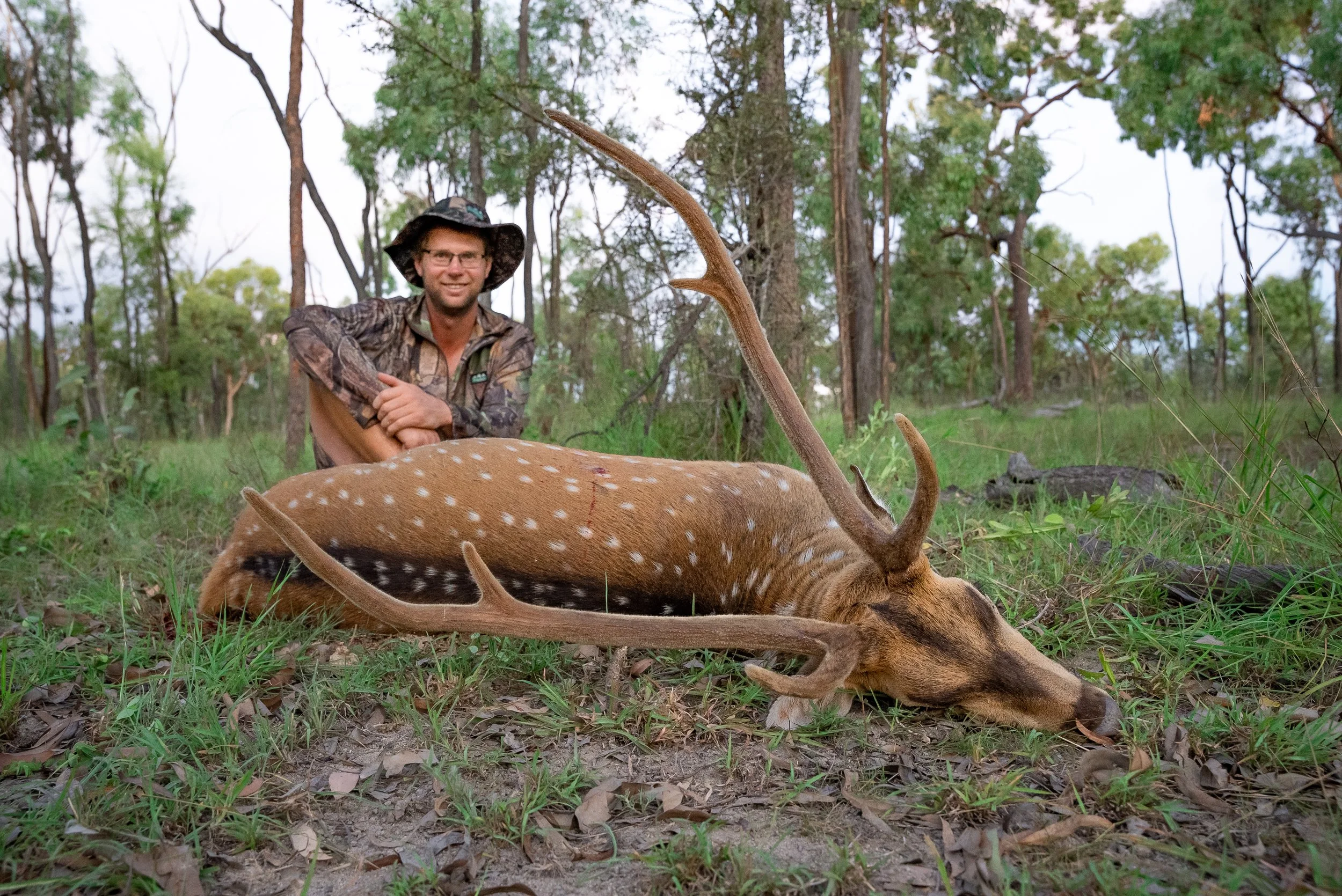 A man in camouflage outdoor clothing and a wide-brimmed hat sits in a forest, smiling, behind a fallen deer.