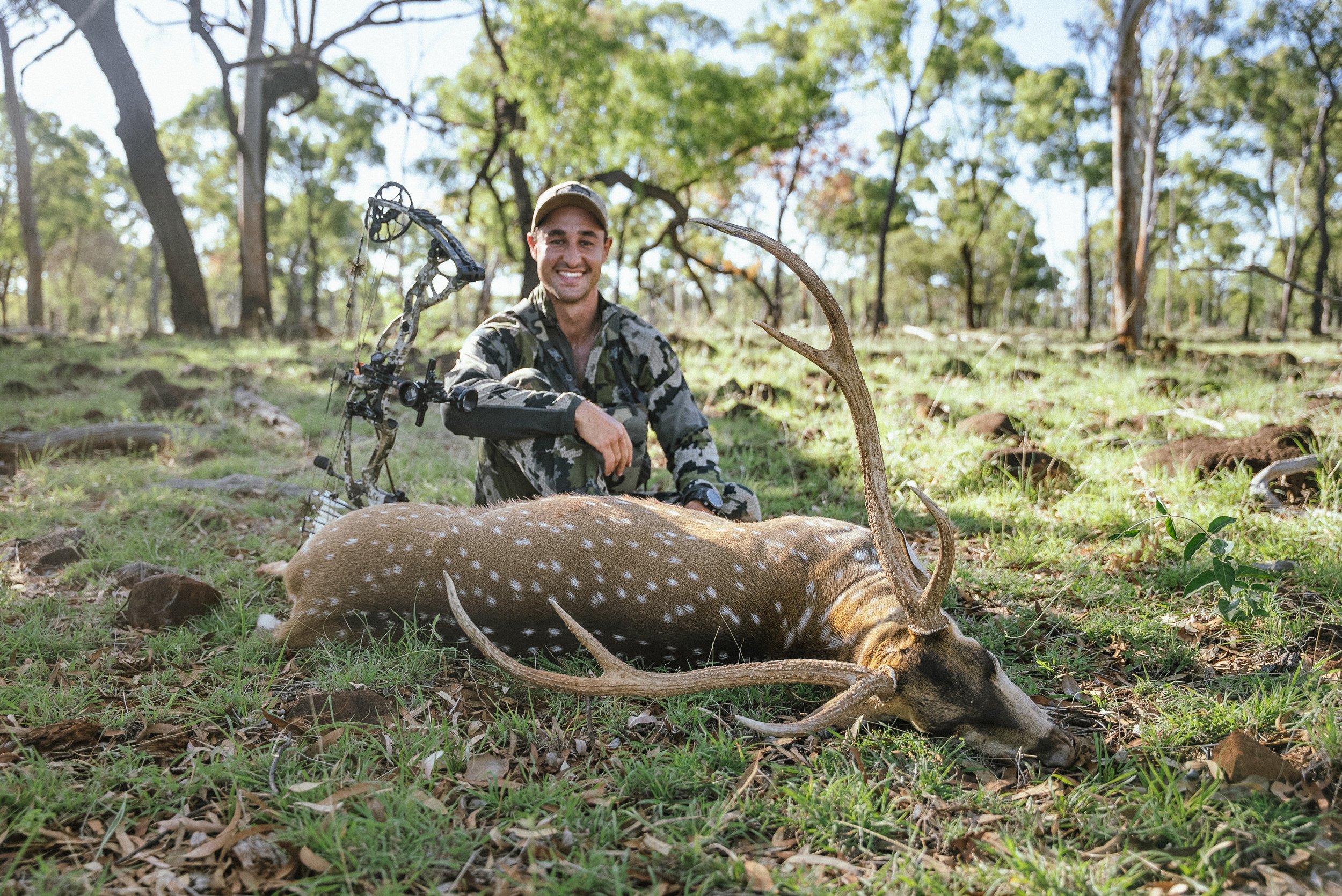 A smiling man dressed in camouflage clothing and a baseball cap kneels on the ground in a forested area, resting his hand on a deer carcass with antlers.