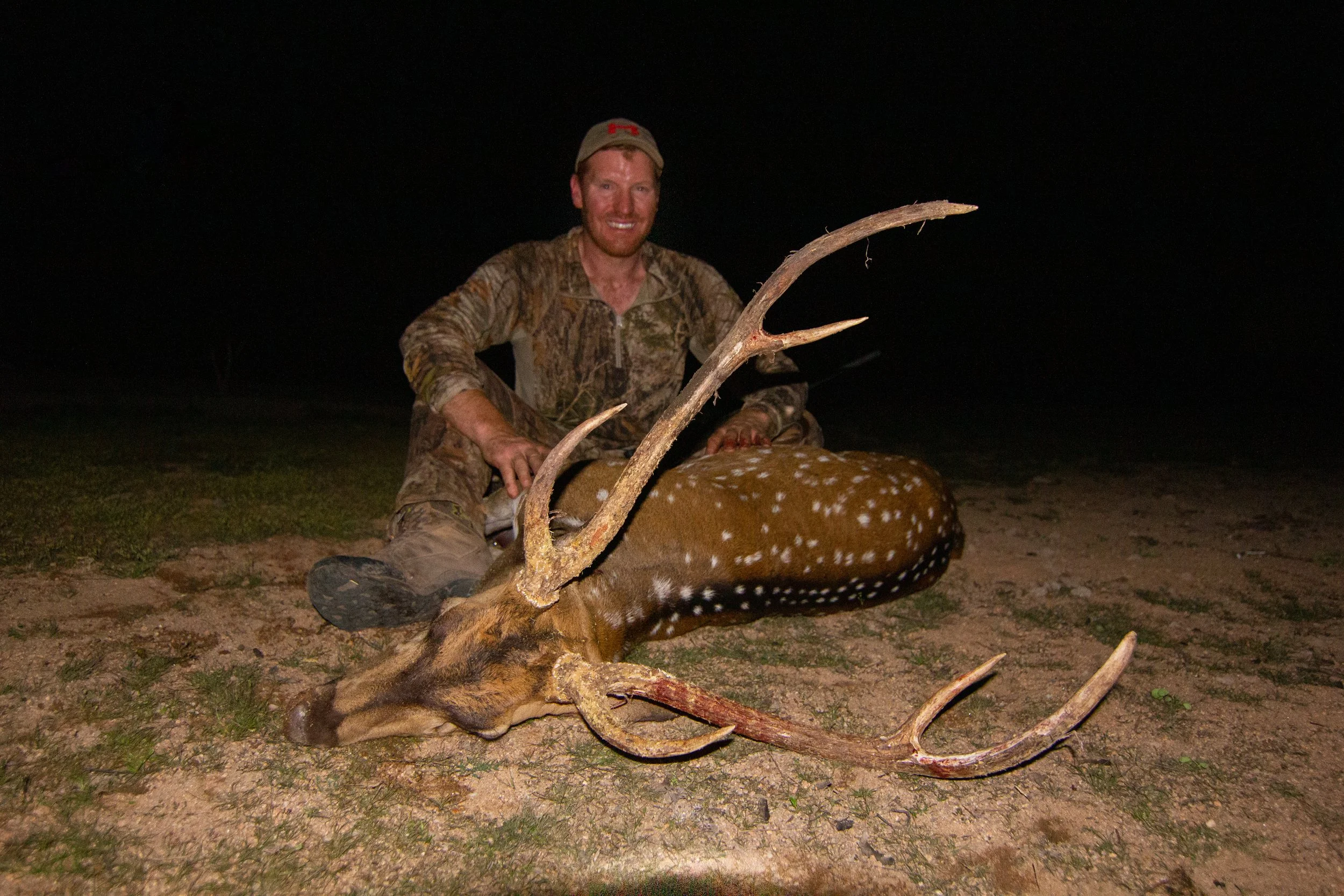 A man in camouflage clothing and a cap kneels at night beside a fallen deer with large antlers, smiling at the camera.