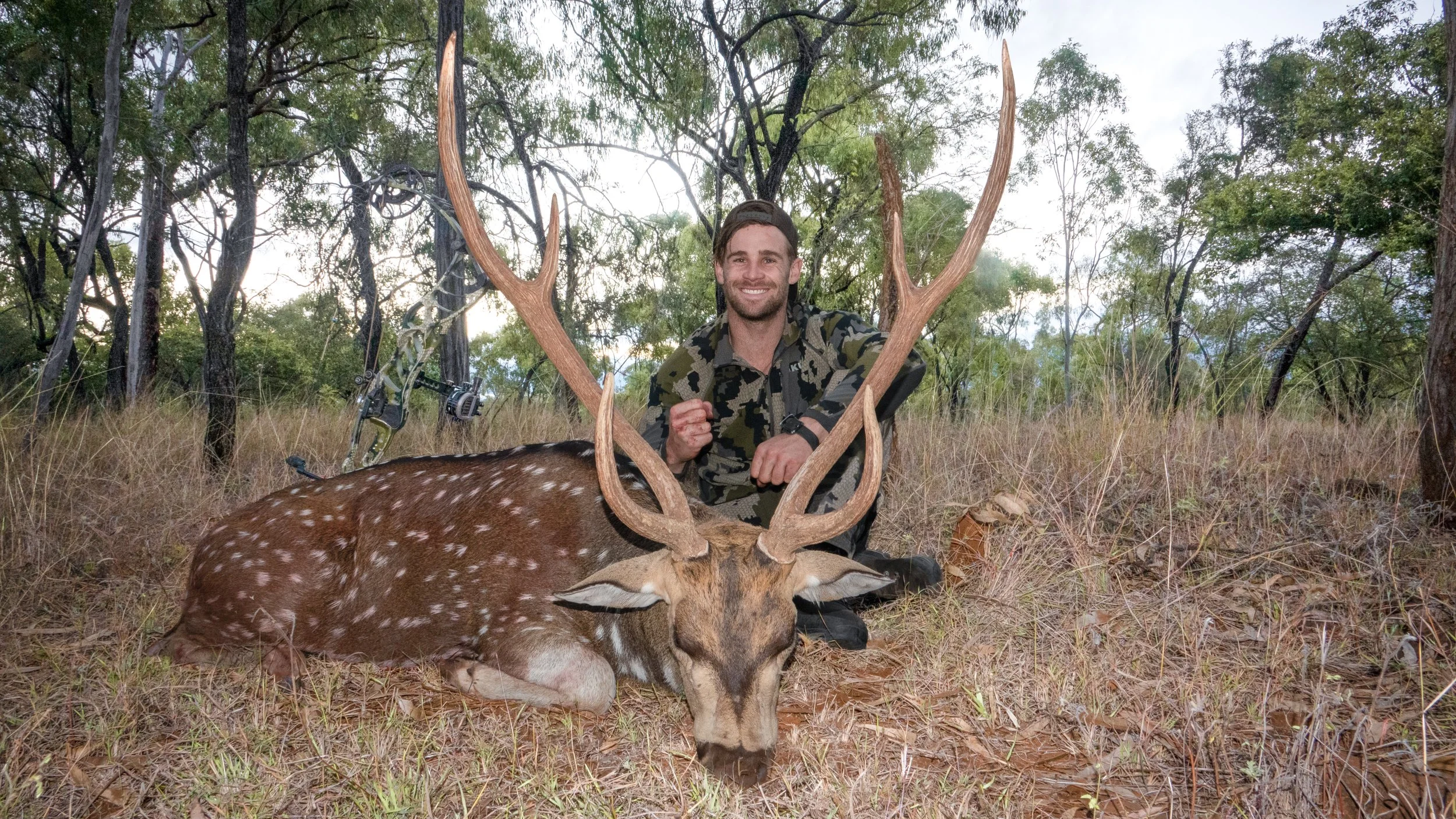 A man smiling while kneeling behind a large deer with antlers lying on the ground, in a wooded area.