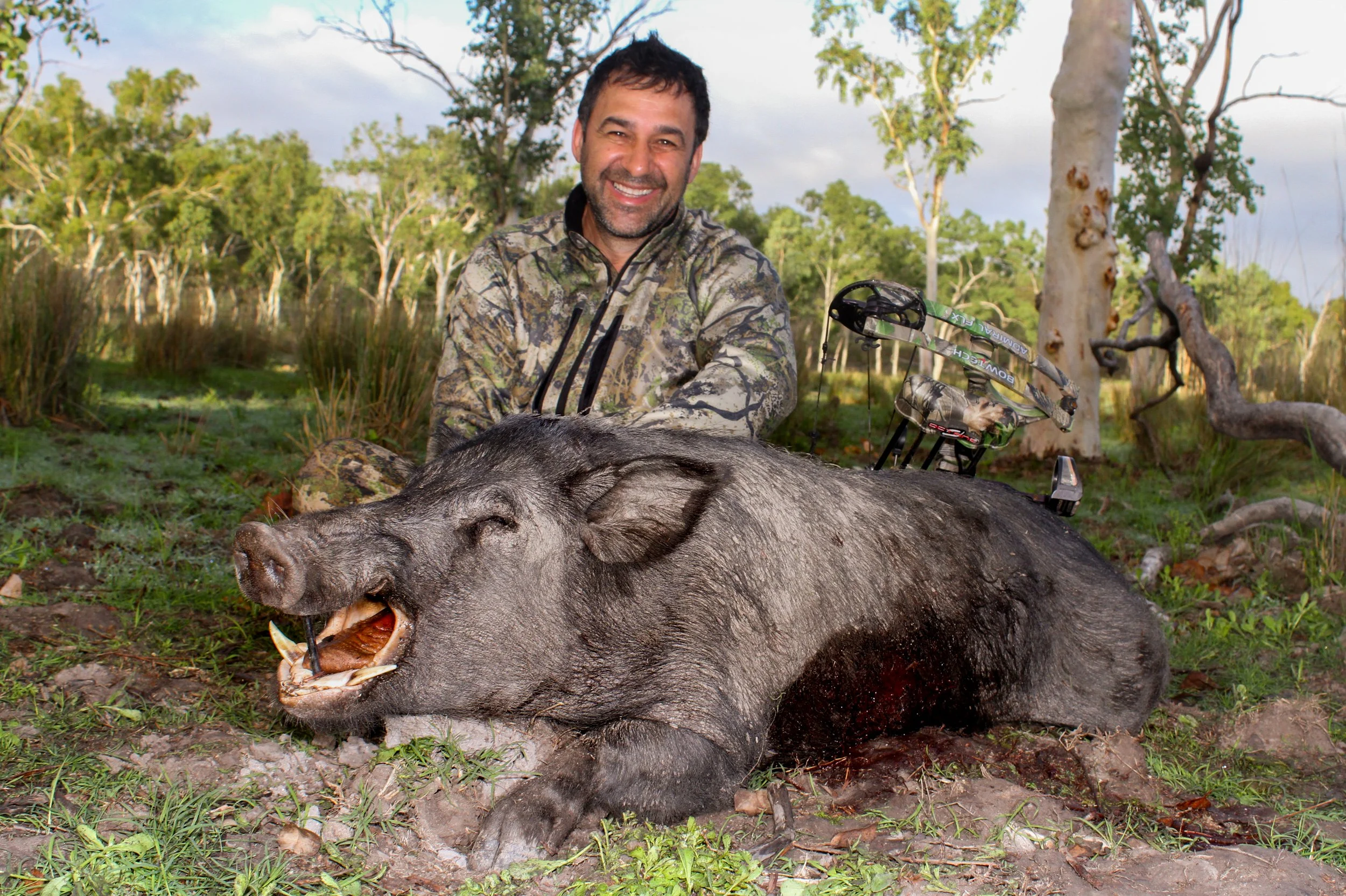 A man in camouflage clothing smiling while sitting behind a large, dead pig on the ground in a forested area.