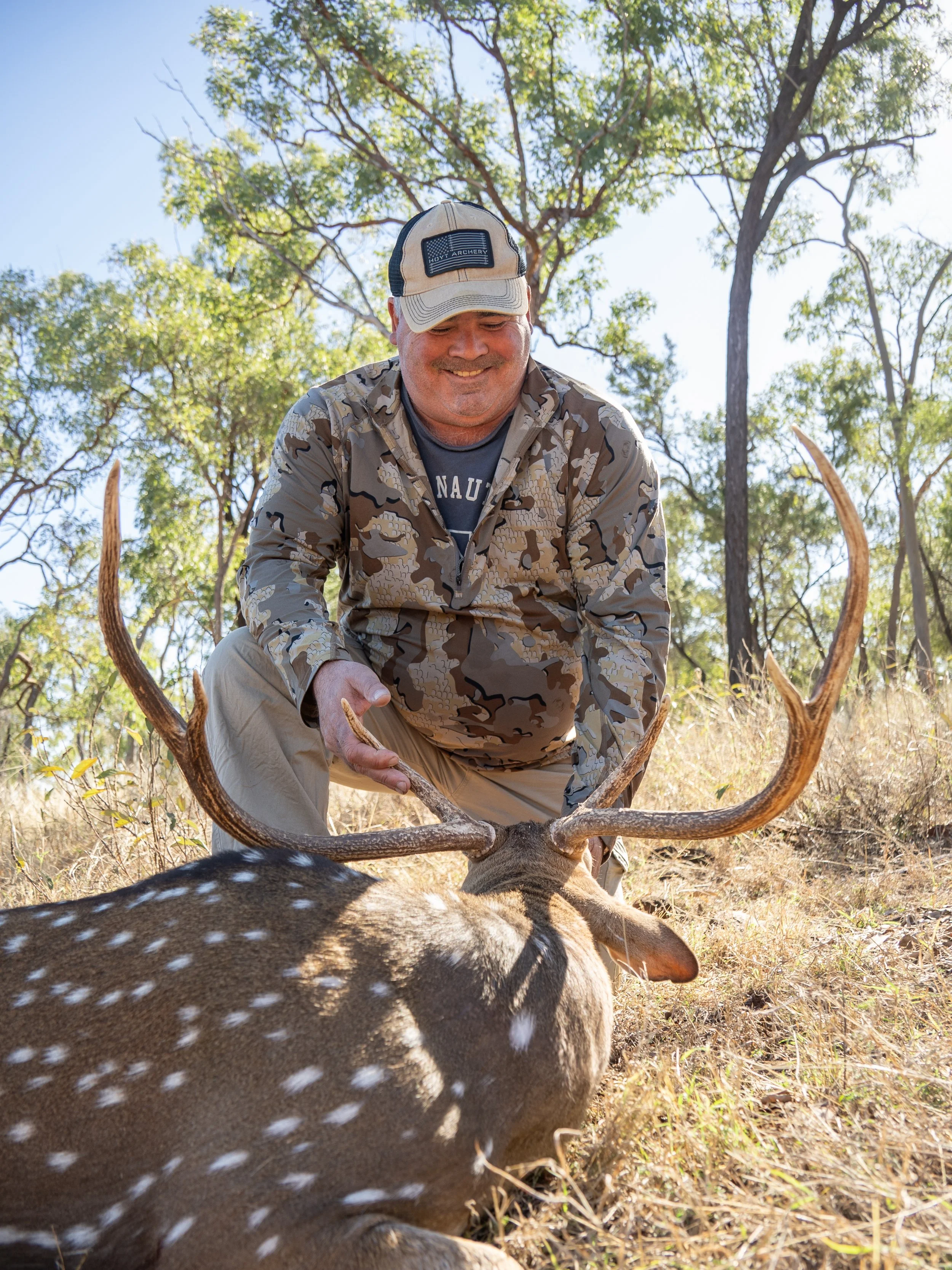 A man in camouflage clothing and a beige cap is kneeling outdoors, holding the antlers of a hunted deer with spotted fur, in a sunny, wooded area with trees and grass.