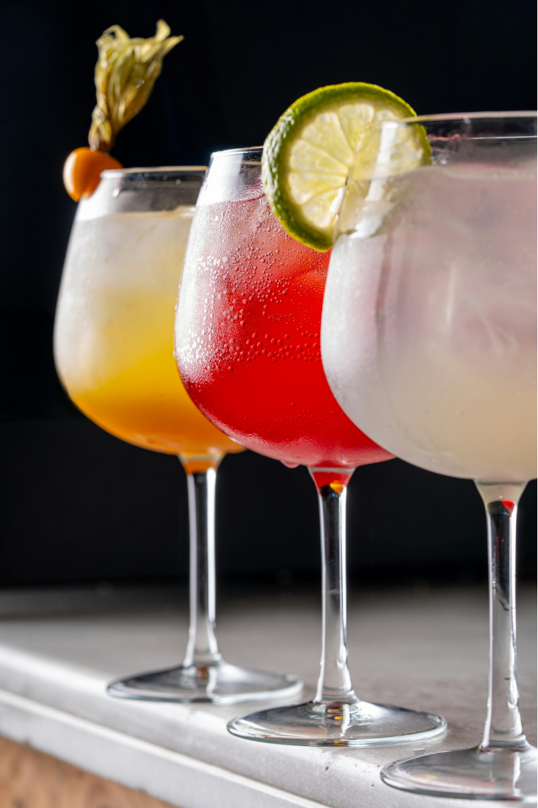 Three colorful mocktails garnished with lime, pineapple, and cherry on a gray surface against a black background.