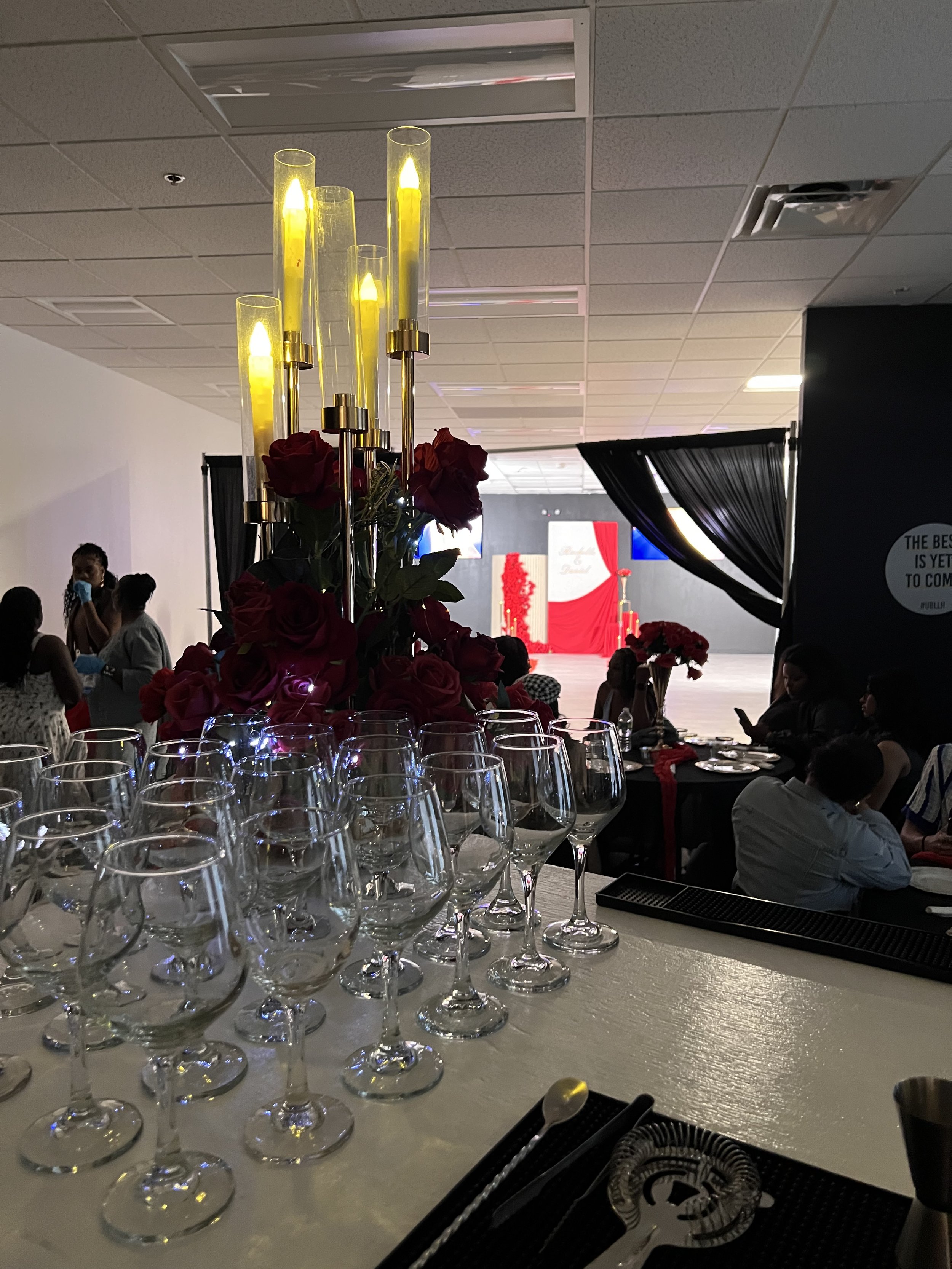 A decorative centerpiece with yellow candles in tall glass holders and red roses surrounded by wine glasses at a banquet table in a large hall with people attending a engagement proposal