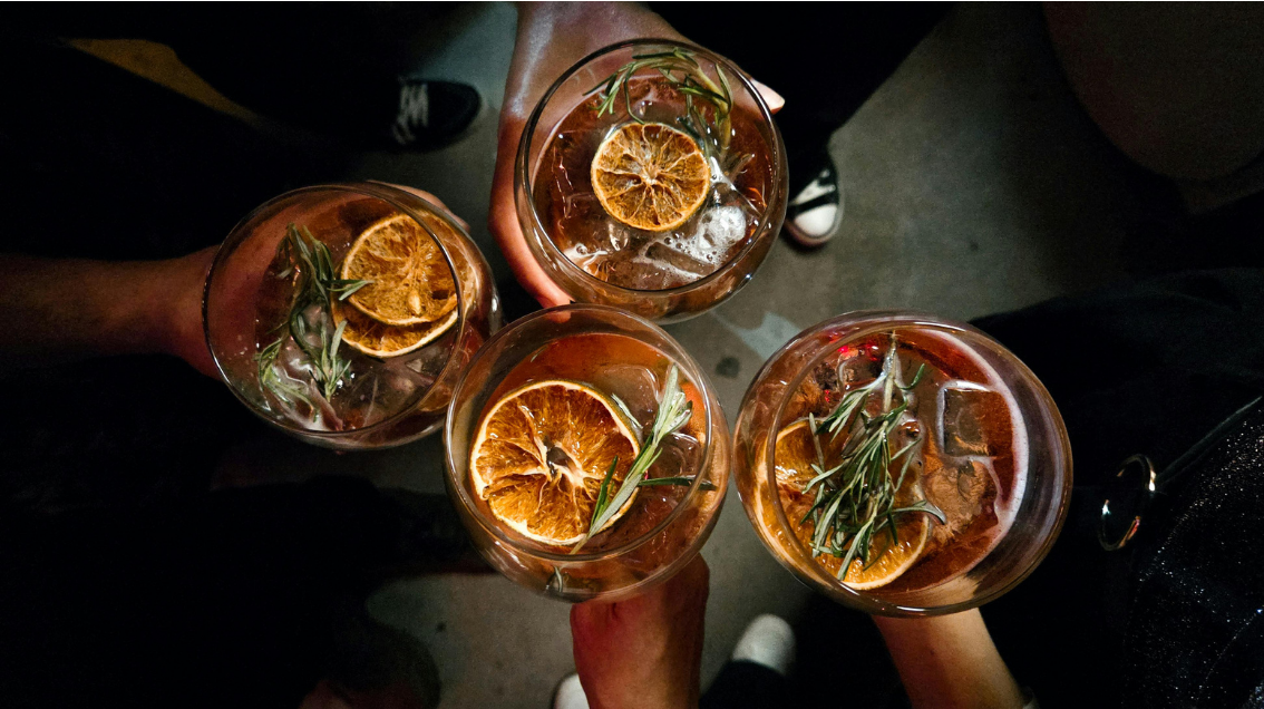 Four glasses of an amber-colored beverage with ice, orange slices, and sprigs of rosemary, being held by four people in a group shot from above.