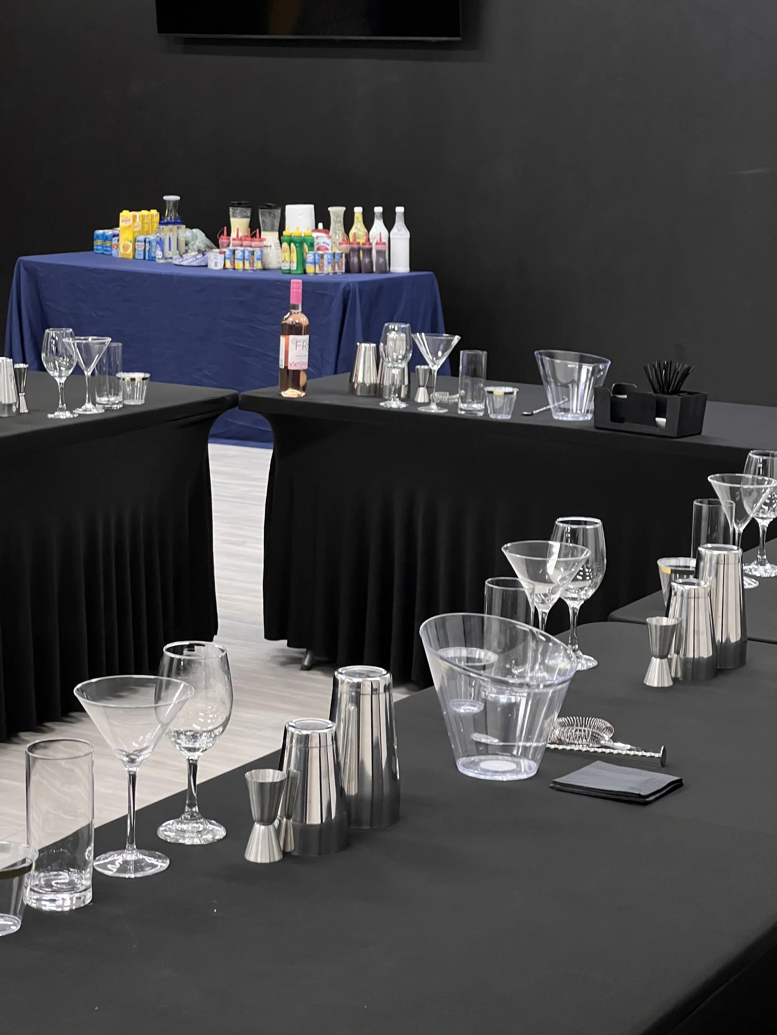 A table setup with various glassware and bar tools, containers of garnishes, and a bottle of rosé wine, in an elegant setting with black tables and a dark background.
