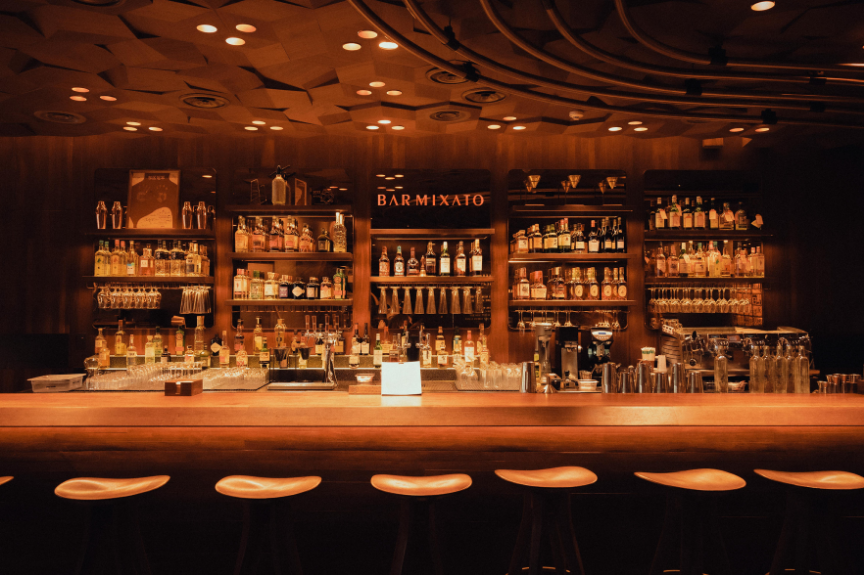 Bar with shelves of liquor bottles, bar tools, and glasses behind a wooden counter with bar stools.