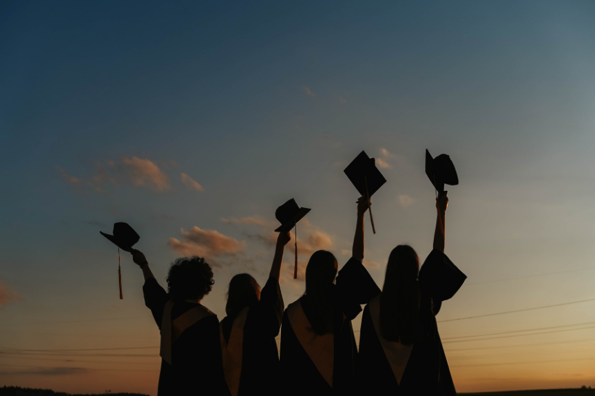 Silhouettes of five graduates in caps and gowns celebrating outdoors at sunset, throwing their caps into the air.