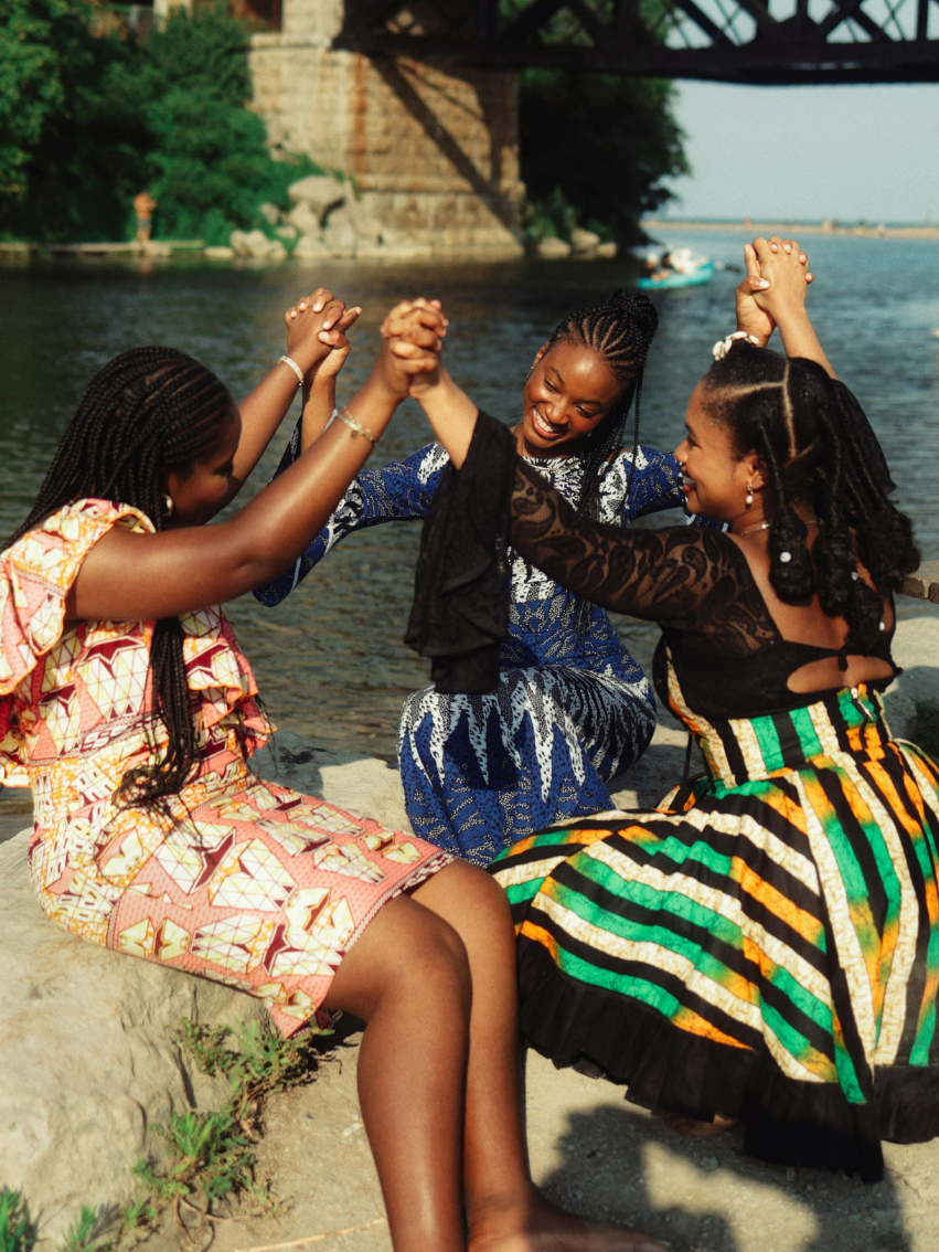 Three women in colorful dresses are sitting by a river, holding hands and smiling, with a bridge and boat in the background.